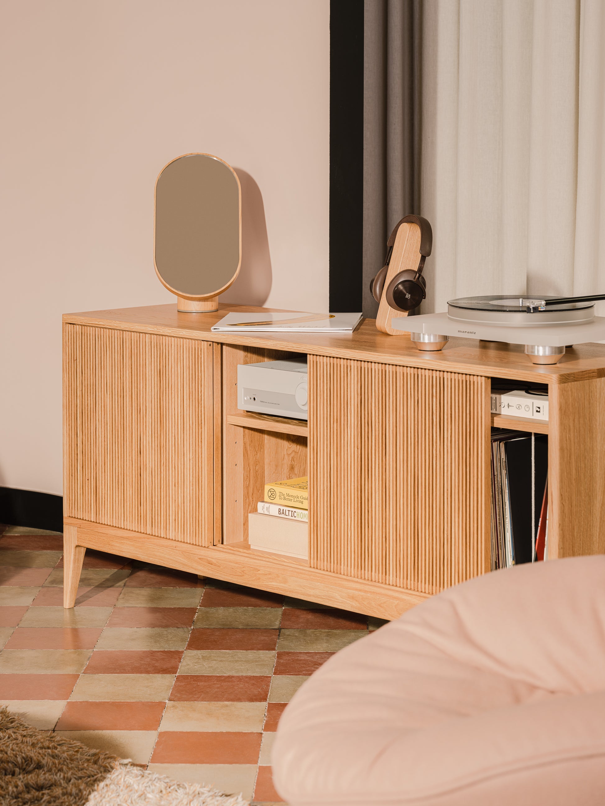 Wooden sideboard with a mirror, headphones, turntable, books, and albums, on checkered floor with curtains.