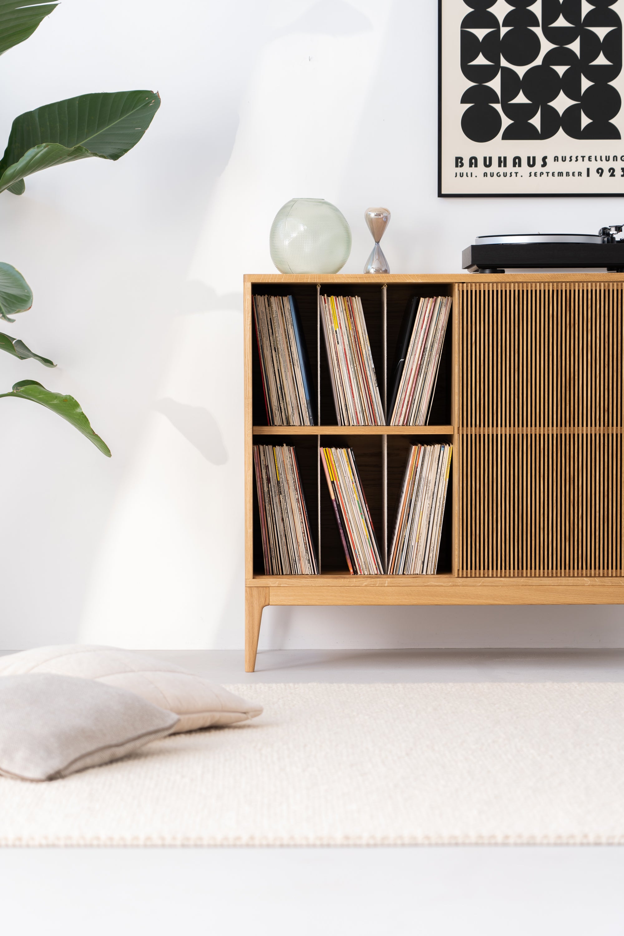 Minimalist room with a wooden record shelf, a vinyl player, a plant, and a Bauhaus poster. A cushion rests on a light rug.