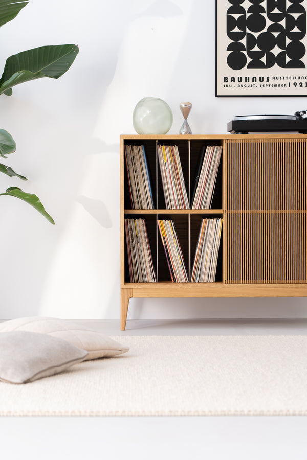 Minimalist room with a wooden record shelf, a vinyl player, a plant, and a Bauhaus poster. A cushion rests on a light rug.