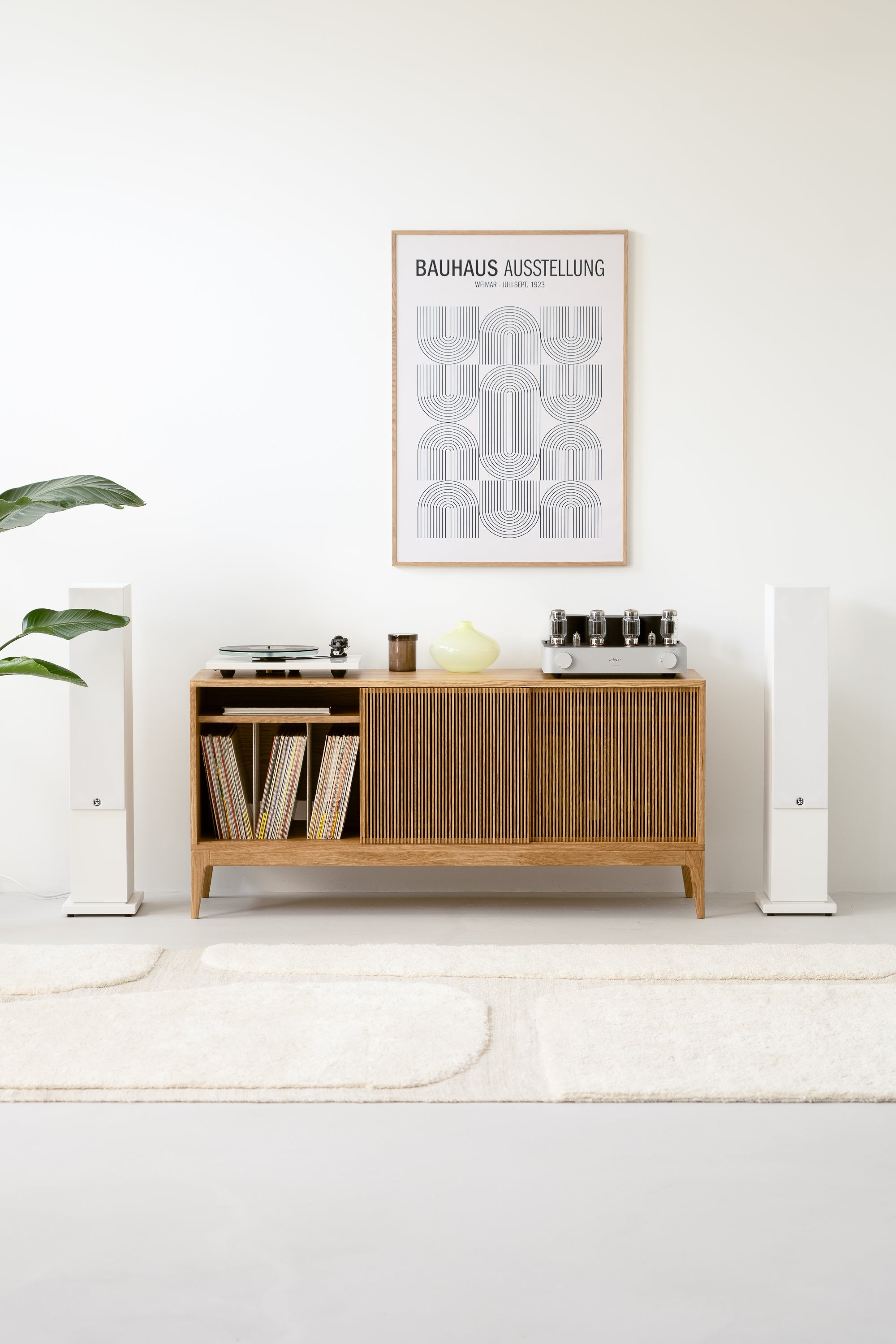 Minimalist living room with a wooden sideboard, Bauhaus poster, vinyl records, speakers, and a potted plant.