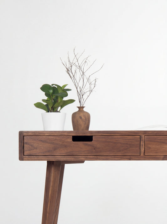 Left corner of the HEIM 140 desk in walnut wood, showing a cut-out drawer grip with a plant and wooden vase above