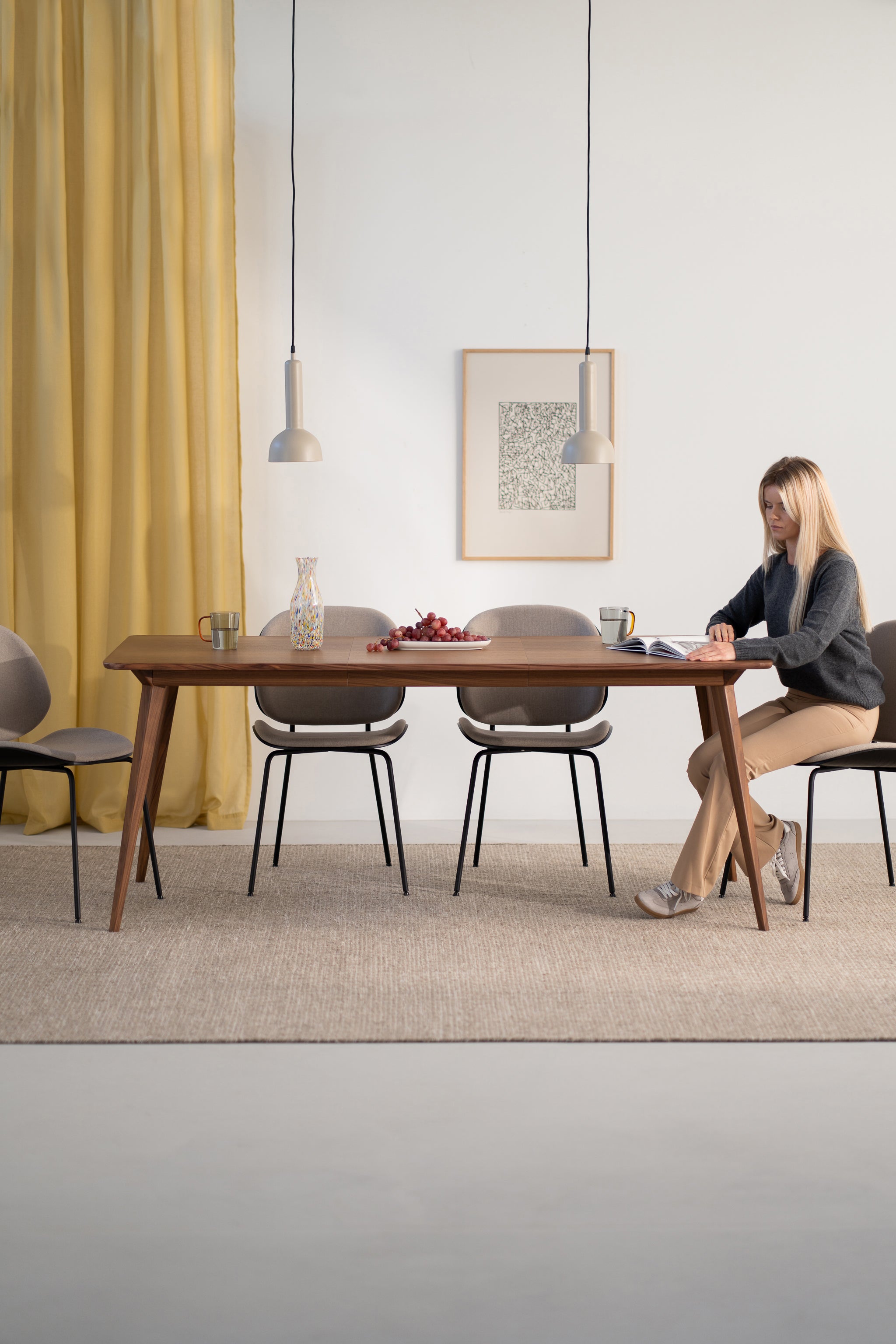 Woman reading at a wooden dining table with chairs, pendant lights, and yellow curtains.