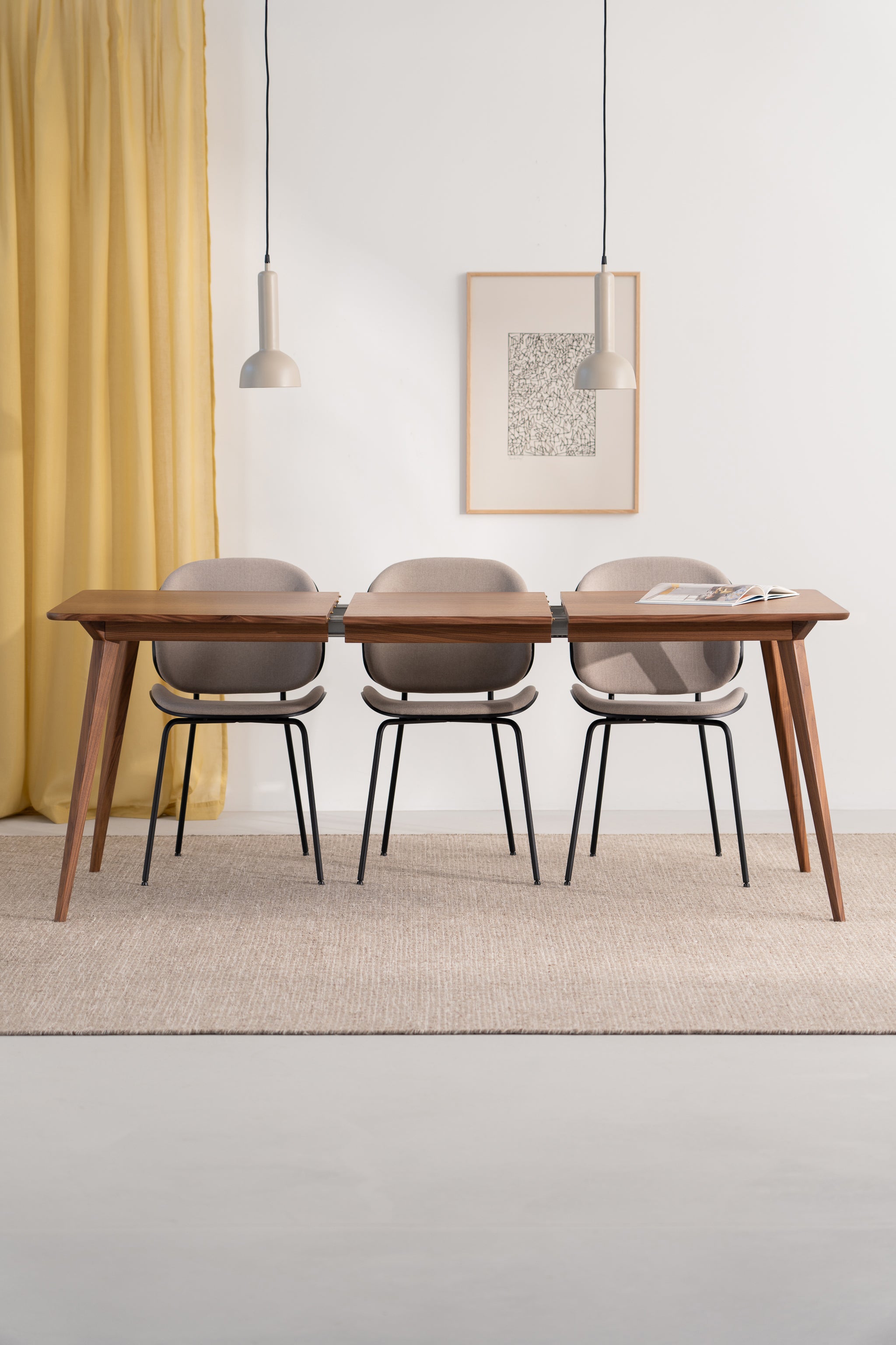 Modern dining set with wooden table, three chairs, pendant lights, and art on a neutral wall with a yellow curtain.