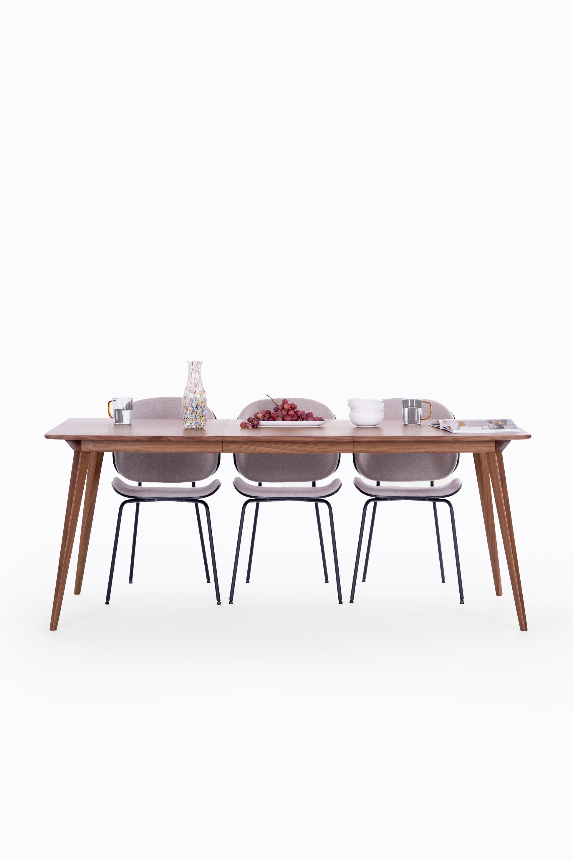 Dining table with three chairs, decorated with grapes, a vase, mugs, and bowls on a clean, white background.