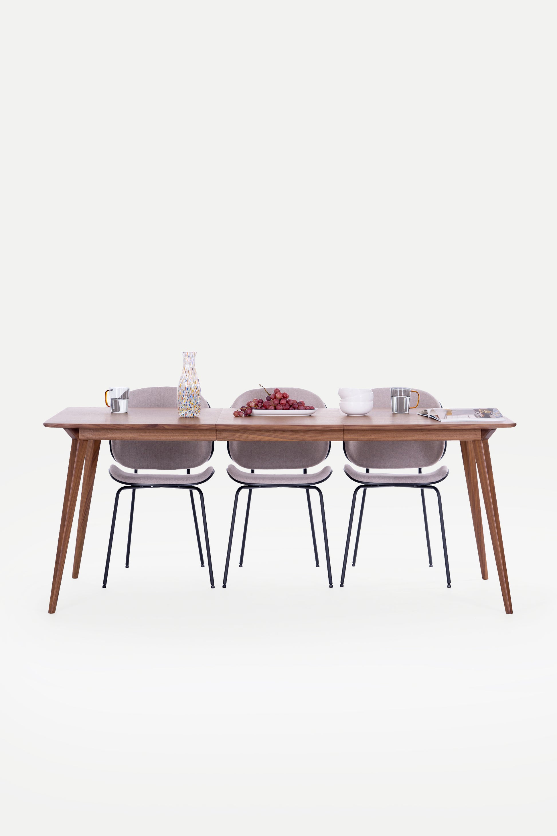 Dining table with three chairs, decorated with grapes, a vase, mugs, and bowls on a clean, white background.