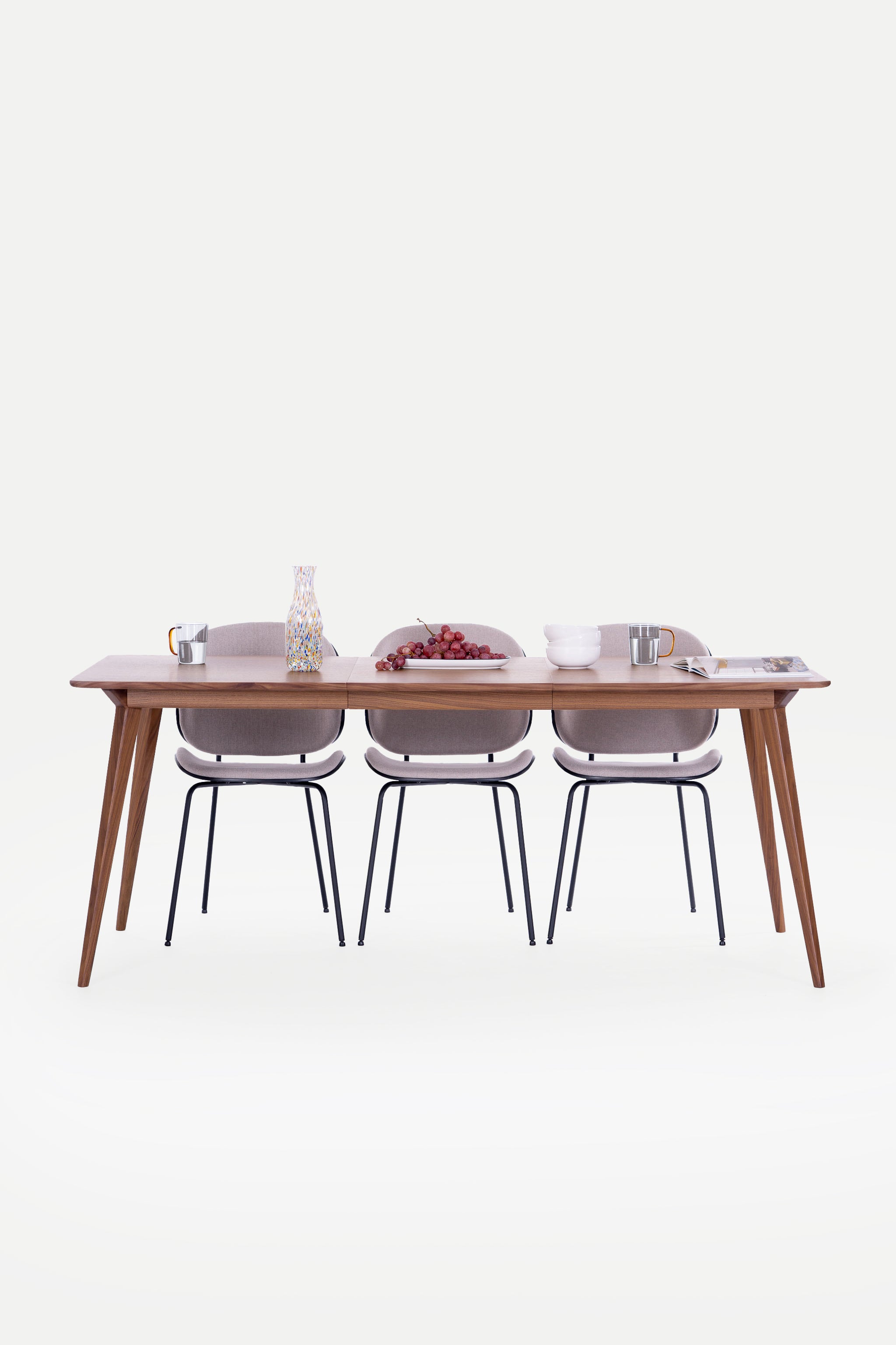 Dining table with three chairs, decorated with grapes, a vase, mugs, and bowls on a clean, white background.