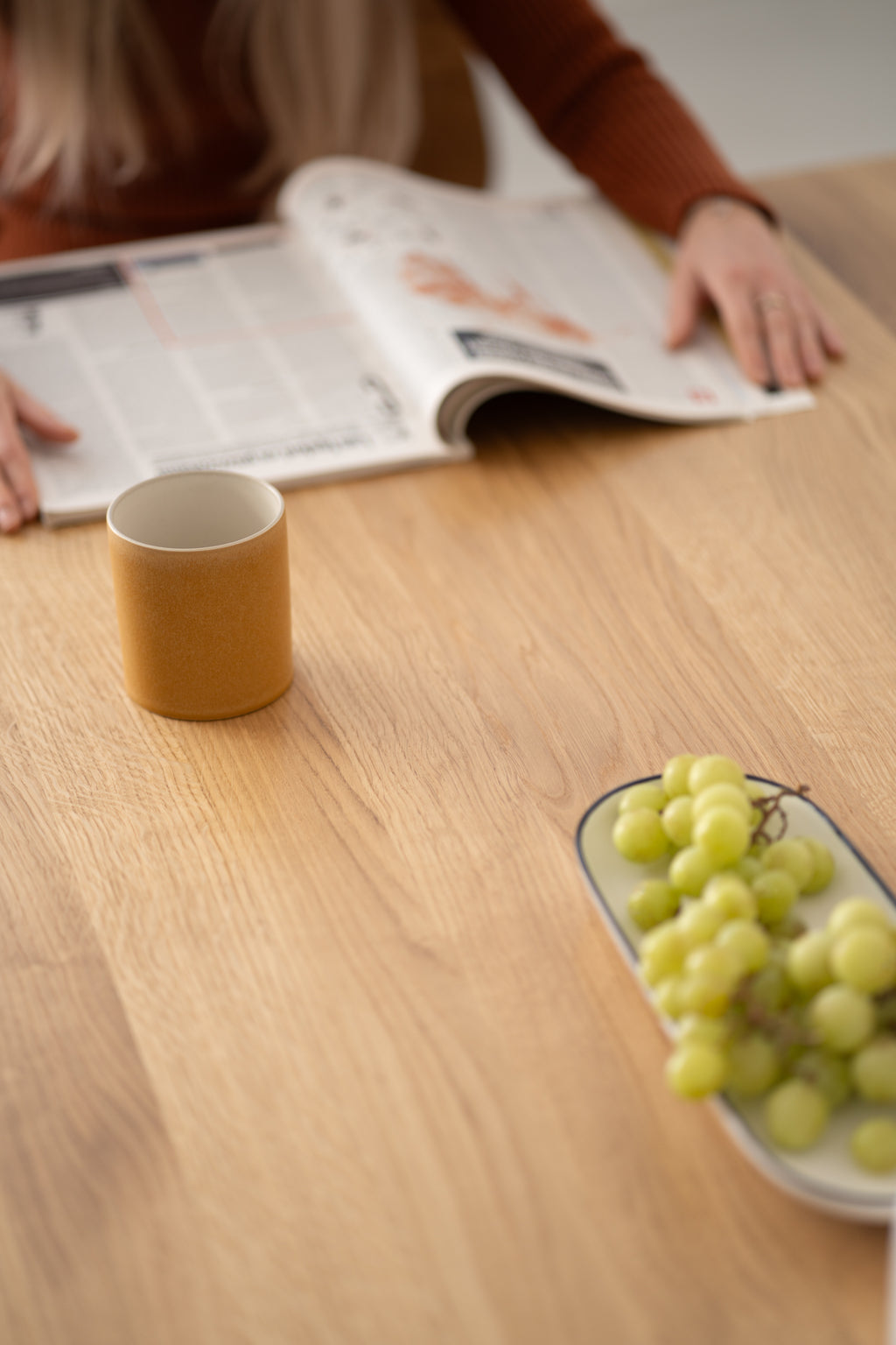 A plate with grapes, a mug, and a person browsing through a magazine on a wooden table.