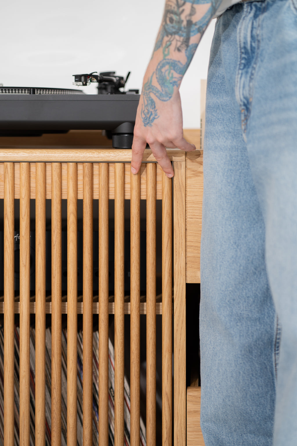 A hand rests on the slatted door's top edge on FUSE X in solid oak, with an amplifier and records visible through the slats