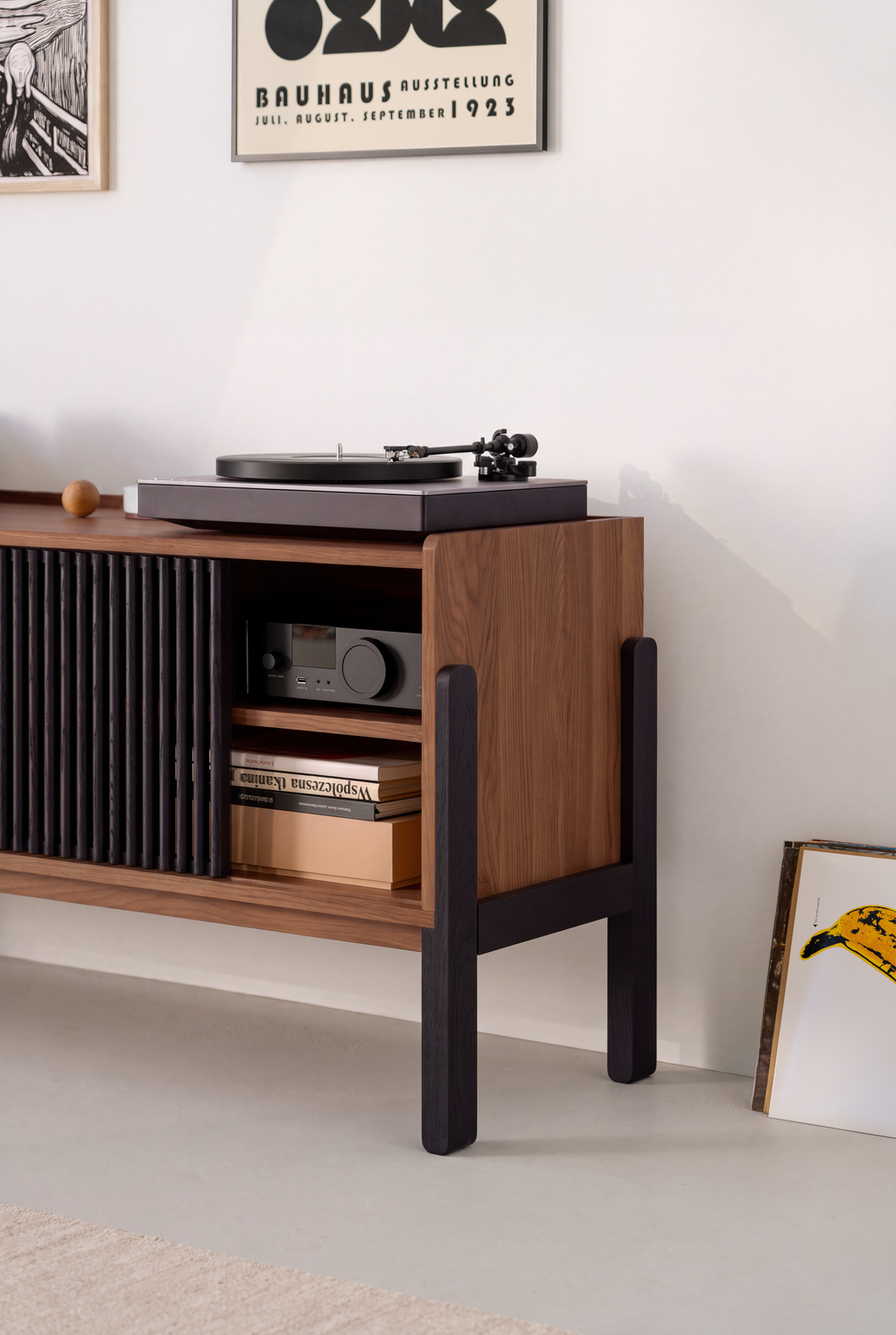 Side view of VIBE in solid walnut on black legs, with a turntable on top and an amplifier above books in the open compartment