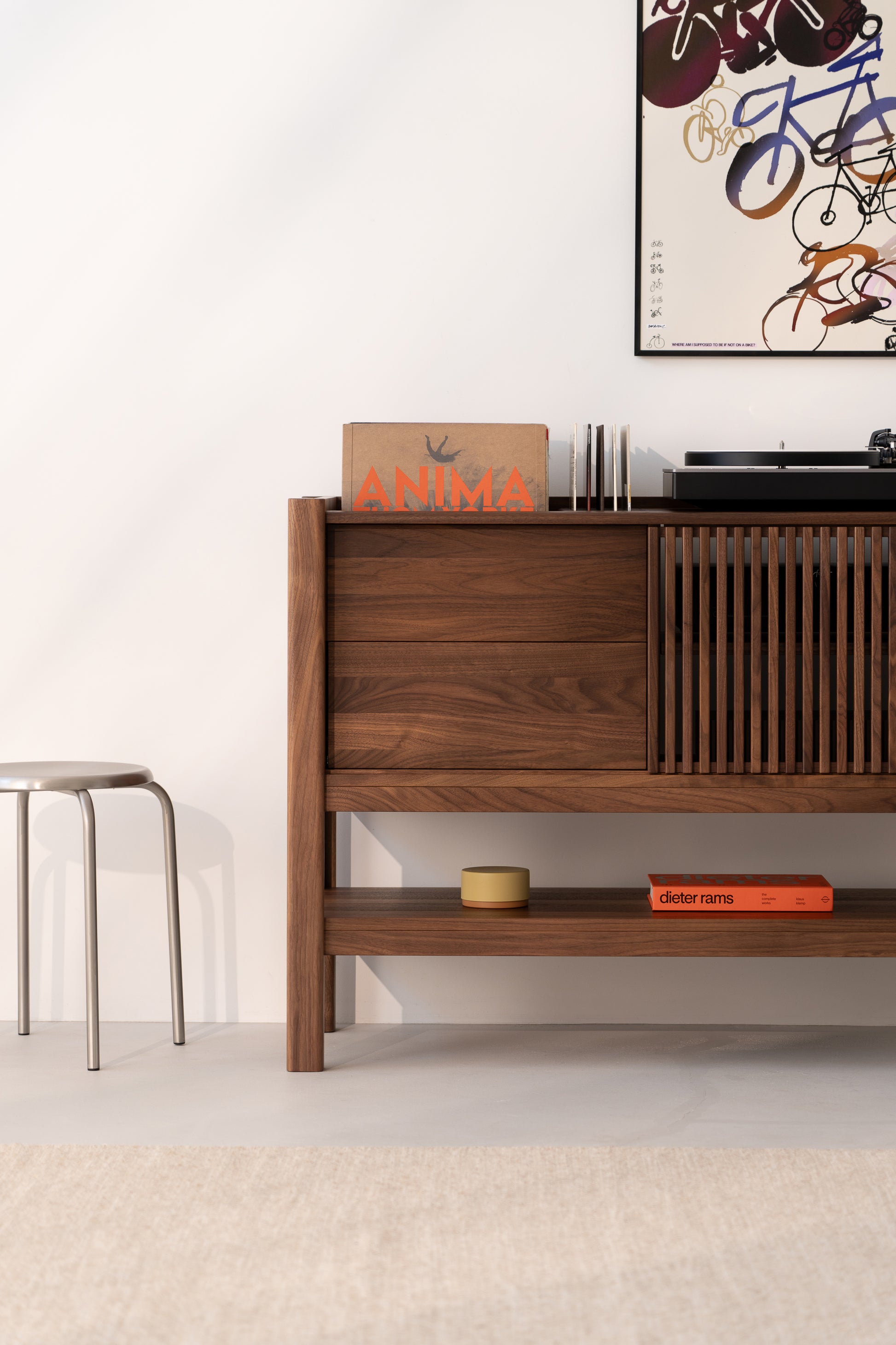 Modern wooden cabinet with books and decor, beside a metal stool, against a light wall with abstract art.