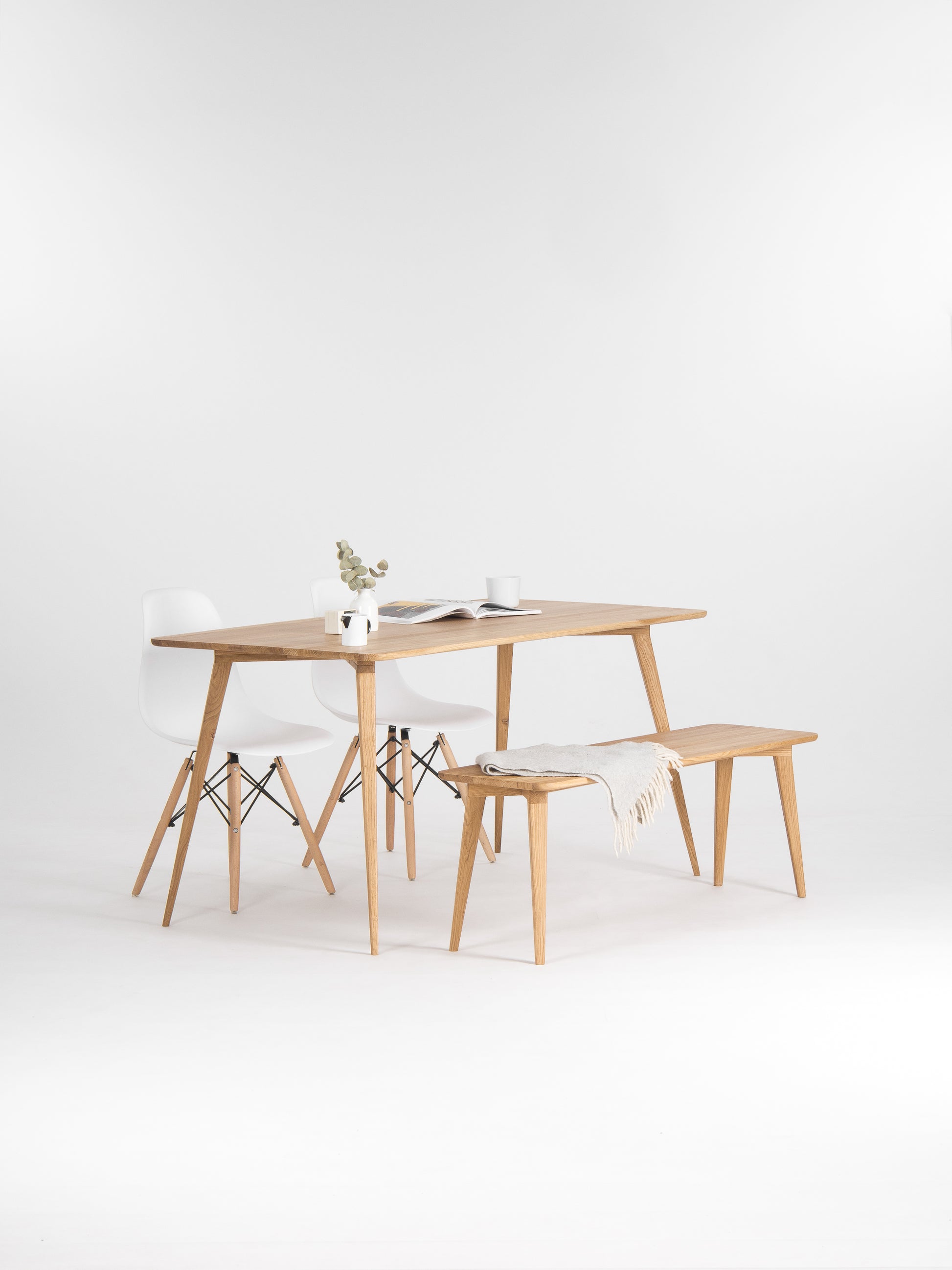 Minimalist dining setup with a wooden table, white chairs, a bench, a vase, and a cup.