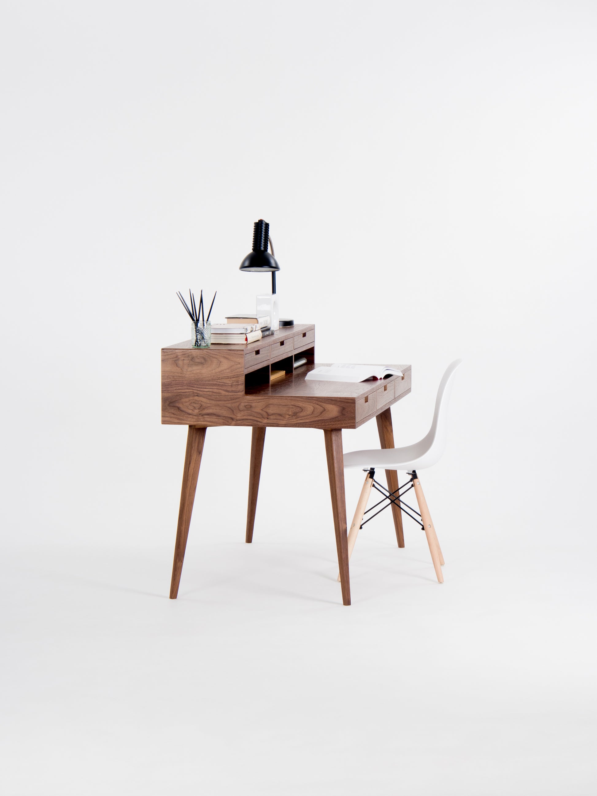 Minimalist wooden desk with a white chair, black lamp, and books against a white background.