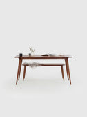 YRKE 8 seater dining table in solid American walnut with bench, front view, gray throw on the backless bench, mugs and a clock