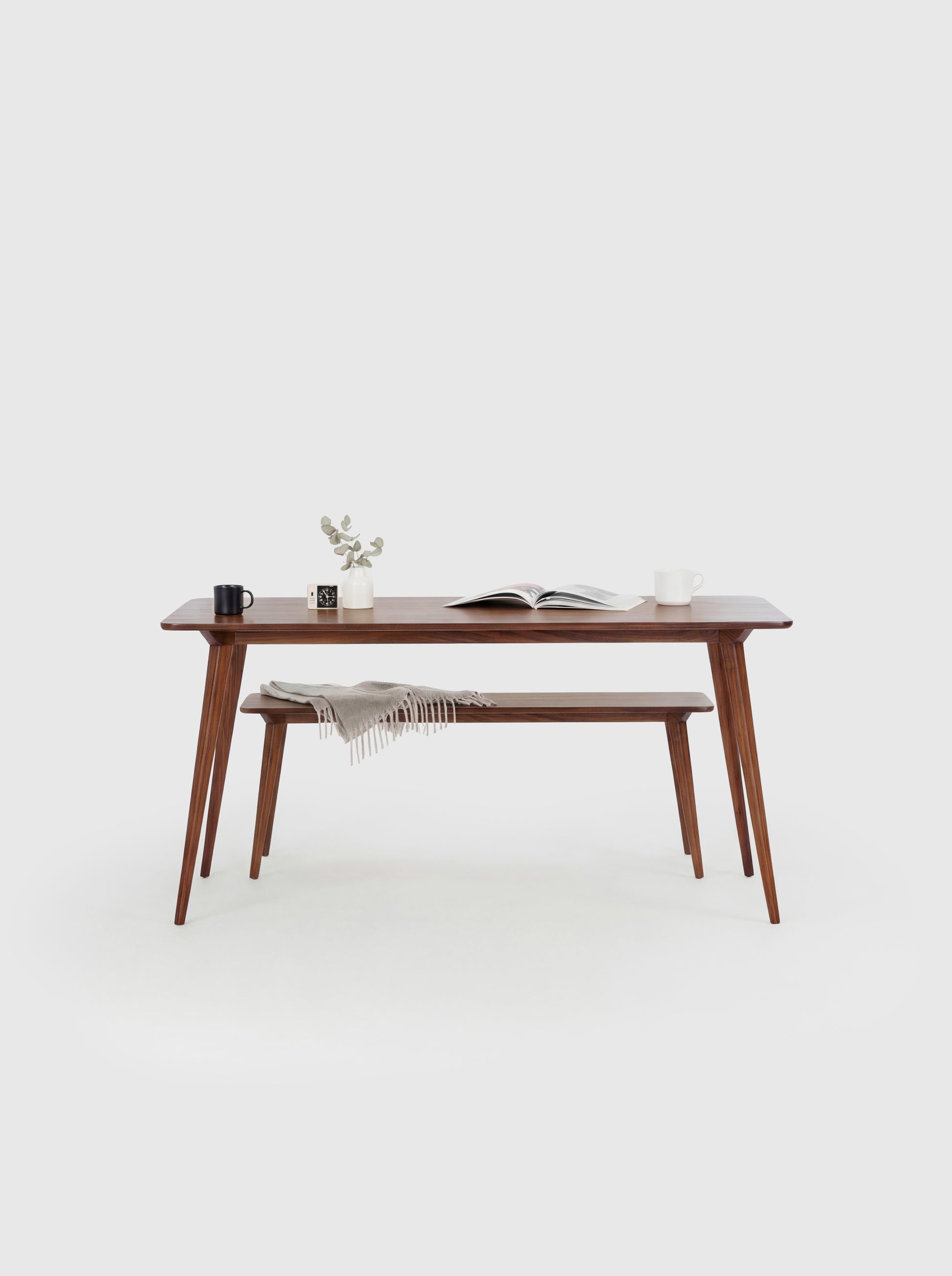 Minimalist wooden table with books, a mug, plant, and bench draped with a blanket in a white room.
