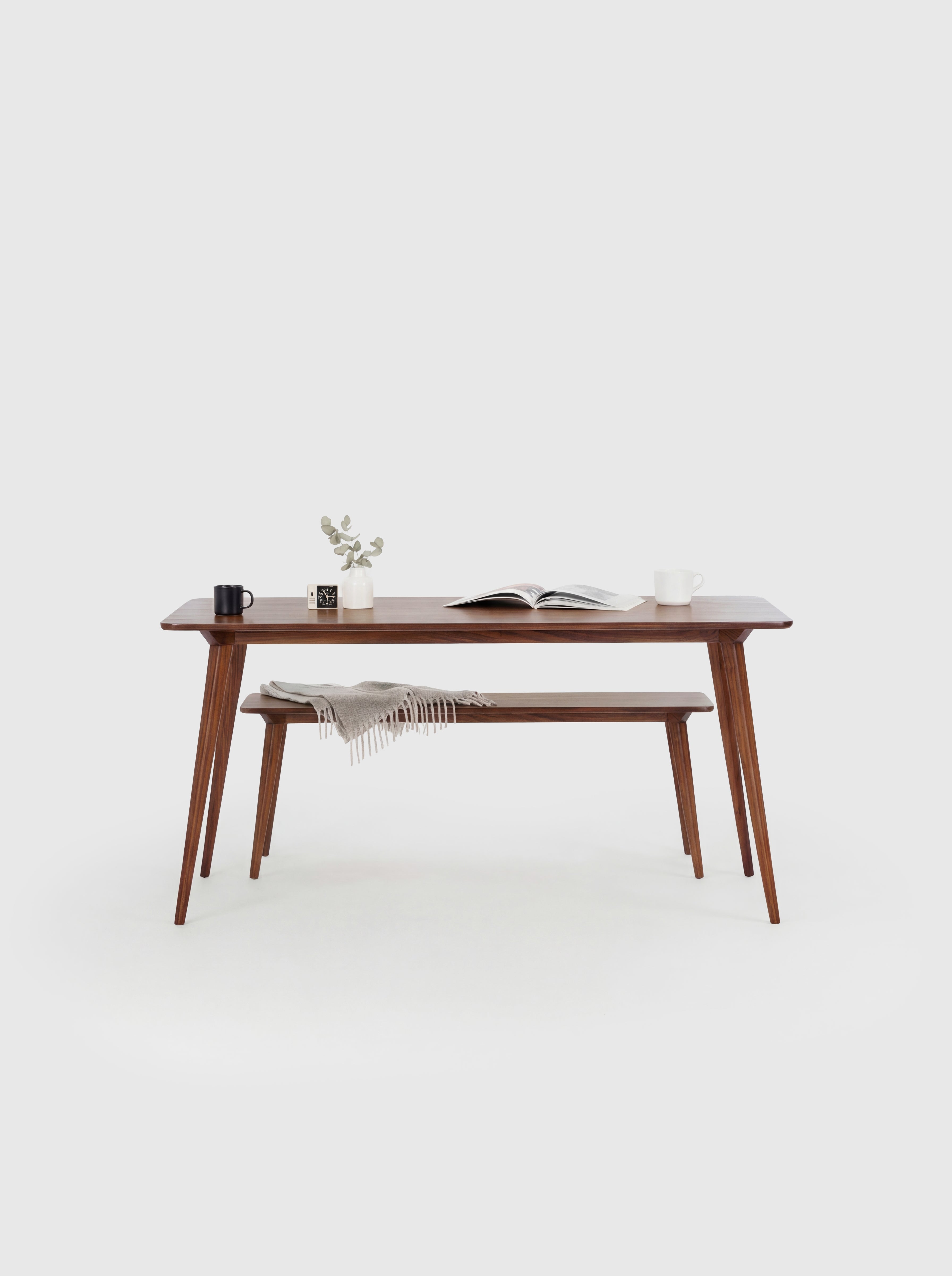 Minimalist wooden table with books, a mug, plant, and bench draped with a blanket in a white room.