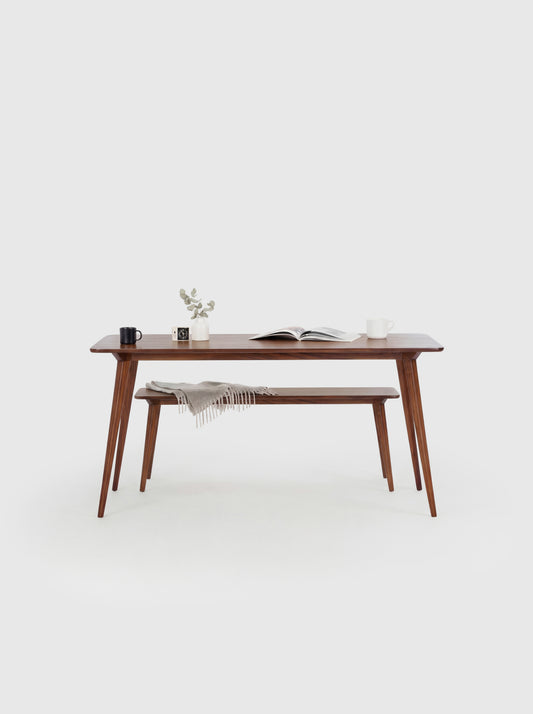 Minimalist wooden table with books, a mug, plant, and bench draped with a blanket in a white room.
