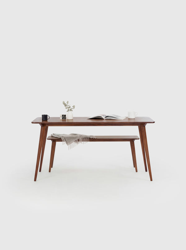 Minimalist wooden table with books, a mug, plant, and bench draped with a blanket in a white room.
