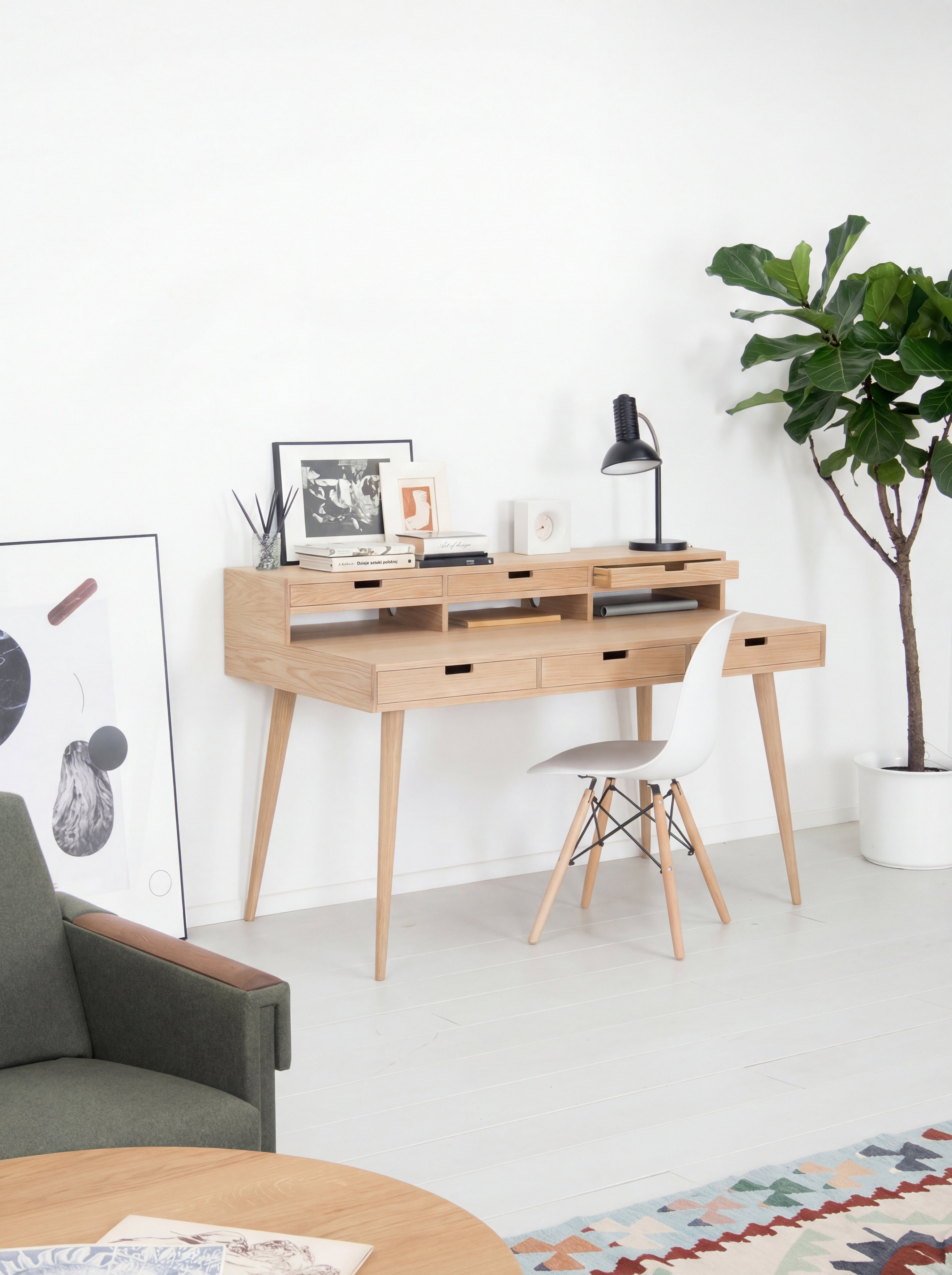 Minimalist workspace with wooden desk, white chair, lamp, potted plant, and wall art in a bright room.