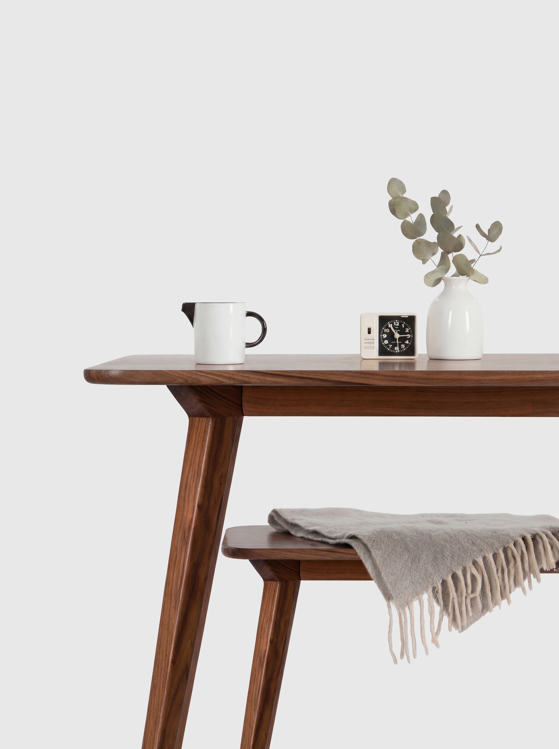 Wooden table with a cup, clock, vase with leaves, and a folded blanket. Minimalist design.