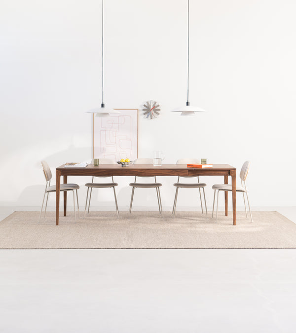 A minimalist dining area with a wooden table, six chairs, two pendant lights, and decor on a light rug.