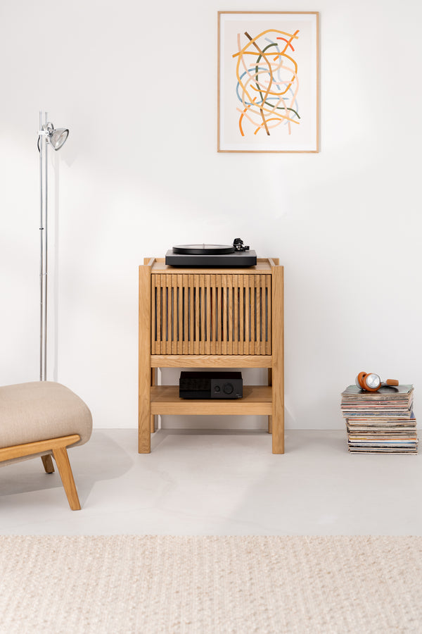 Minimalist living room with a wooden cabinet, turntable, artwork, lamp, and stack of vinyl records.