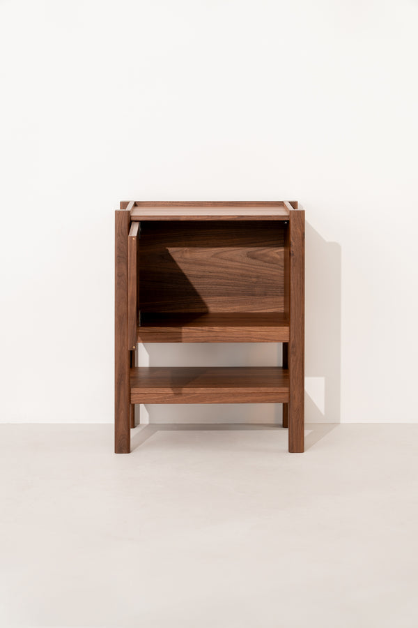 Wooden side table with a minimalist design, featuring an open shelf and a top compartment against a white wall.