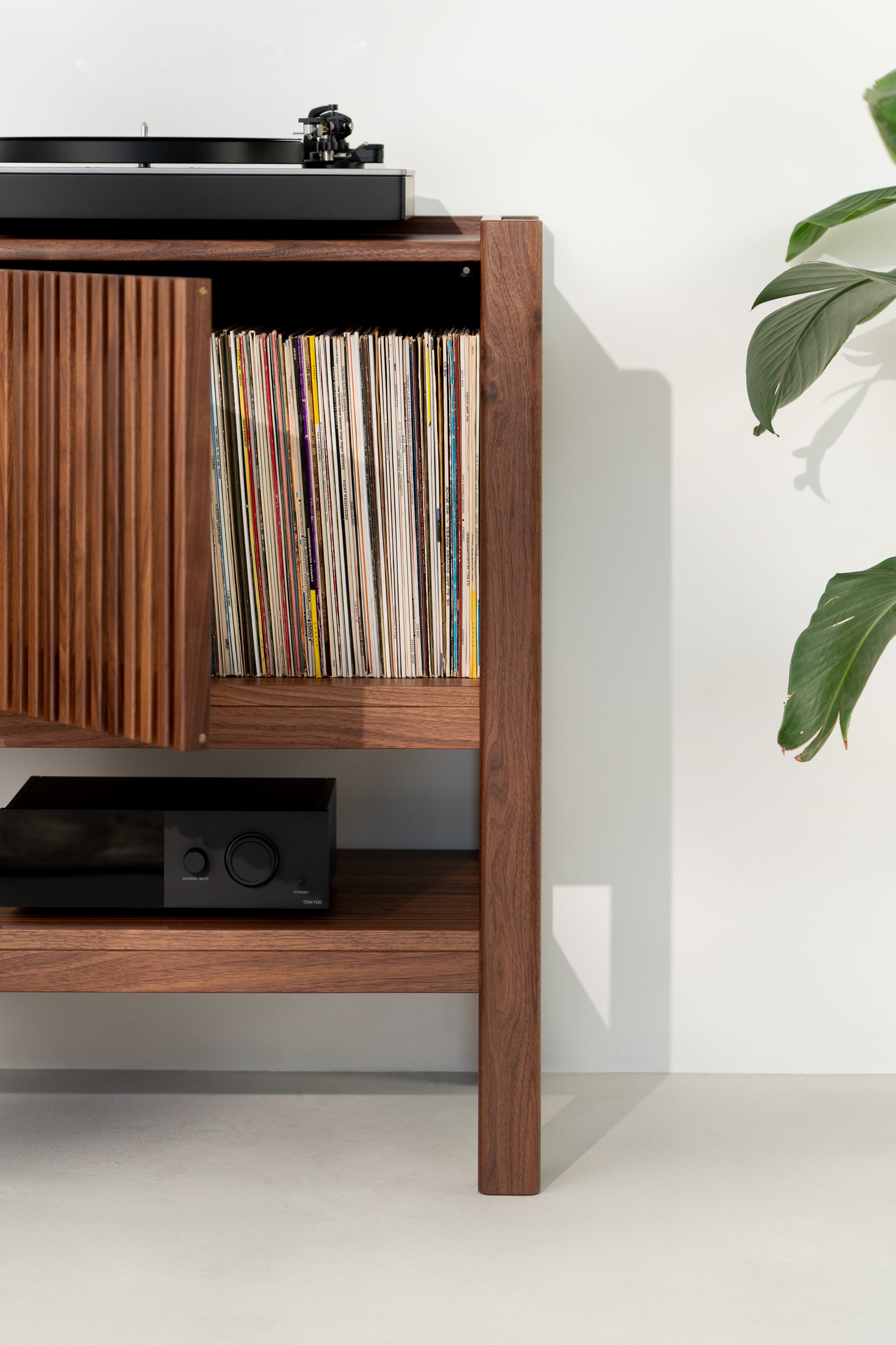 Wood cabinet with vinyl records, a record player on top, and a black amplifier on the shelf below, next to a plant.
