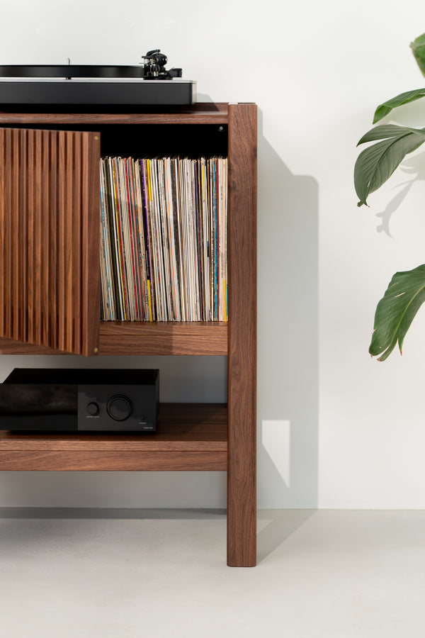 Wood cabinet with vinyl records, a record player on top, and a black amplifier on the shelf below, next to a plant.
