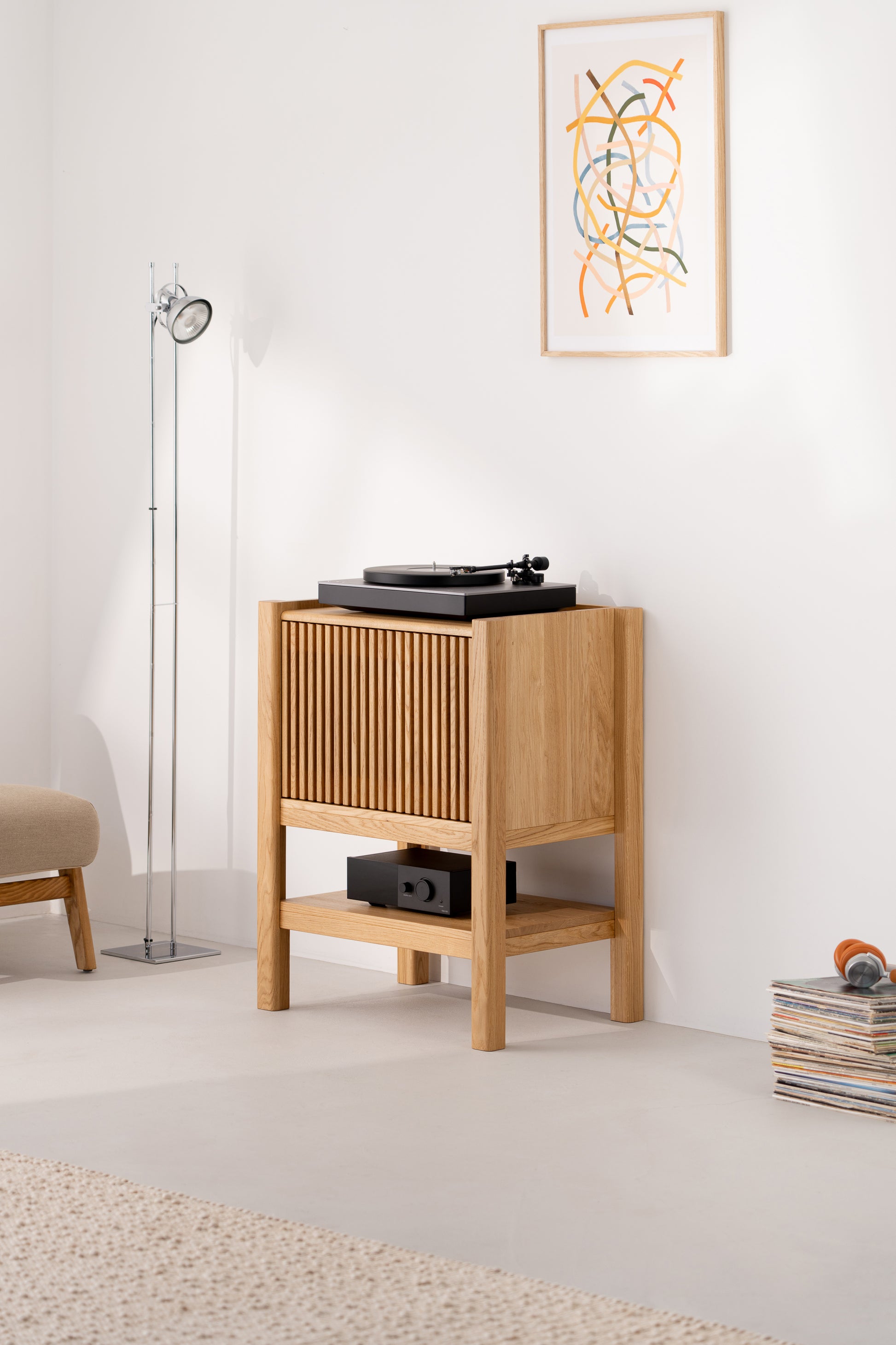 Wooden record player stand with turntable, lamp, abstract art, and stack of vinyl records in a modern room.
