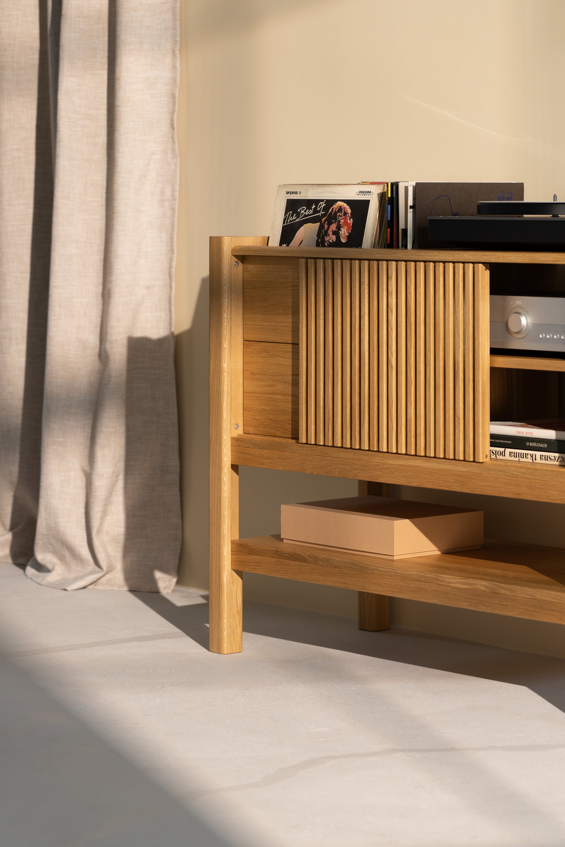  A wooden shelf with vinyl records, a stereo, and a pink box, next to beige curtains.