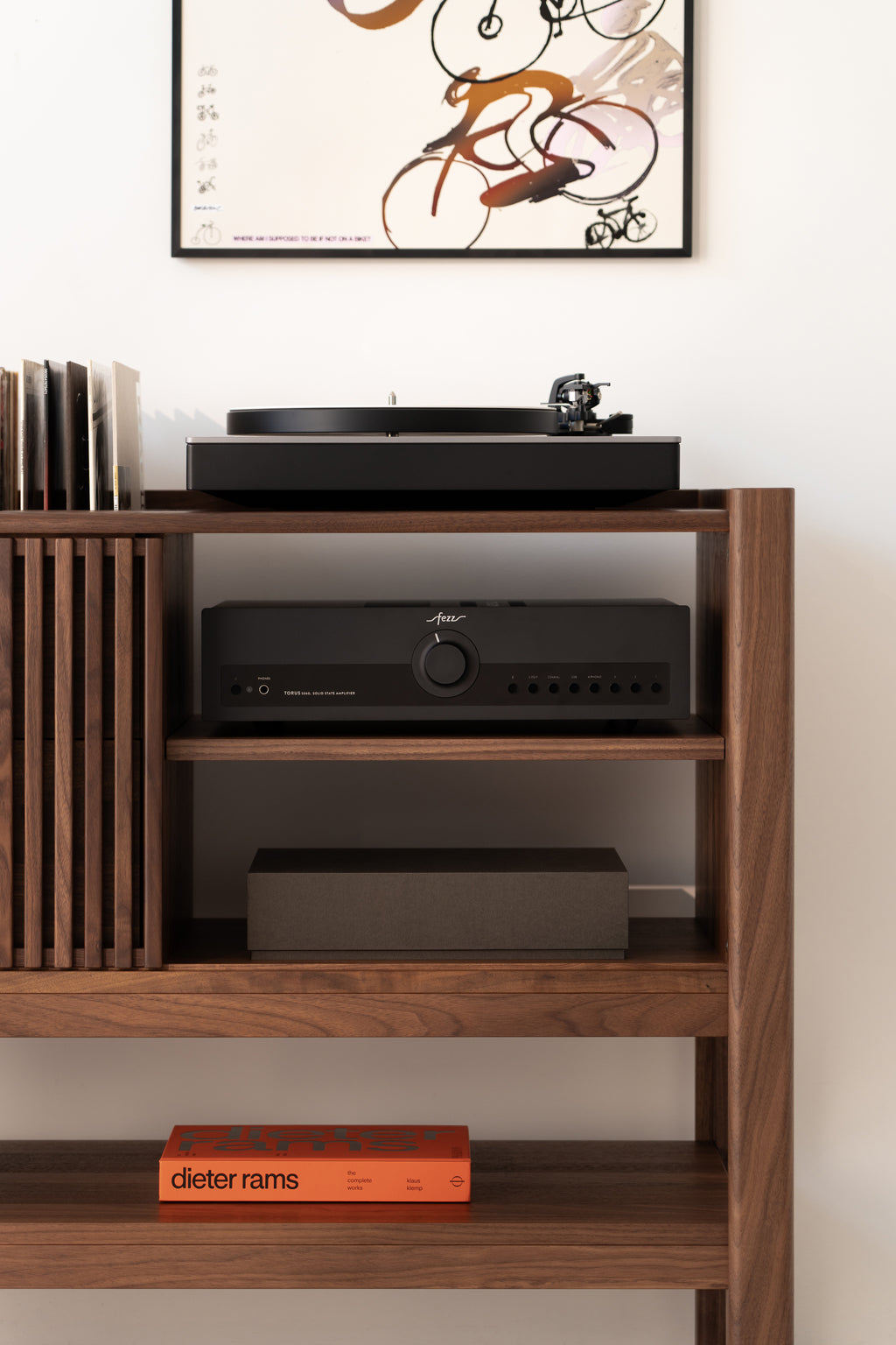 Wooden shelf with a turntable, records, an amplifier, and a book titled "Dieter Rams," below an abstract art print.