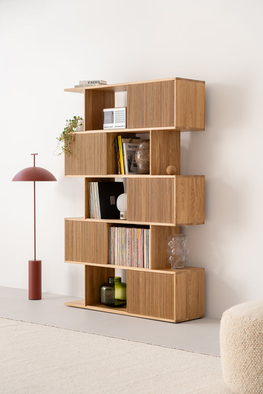 Wooden bookshelf with books, plants, and decor items. Red floor lamp and a textured stool nearby.