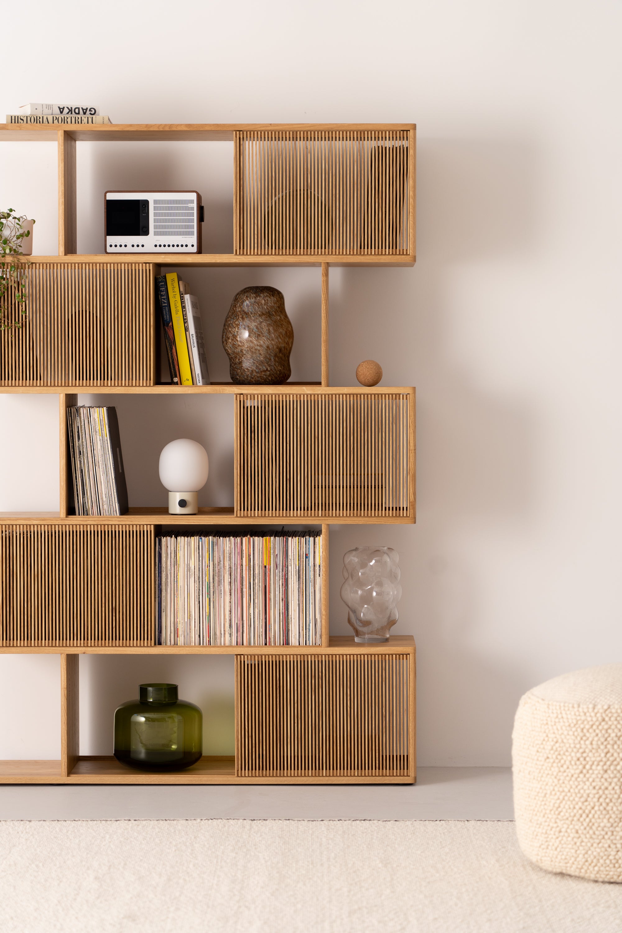 Wooden shelf with books, vases, a radio, lamp, and records. Beige wall and pouf nearby.