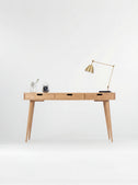 Front view of the HEIM 140 computer desk in oak wood, with three wooden drawer fronts, a brass lamp and alarm clock