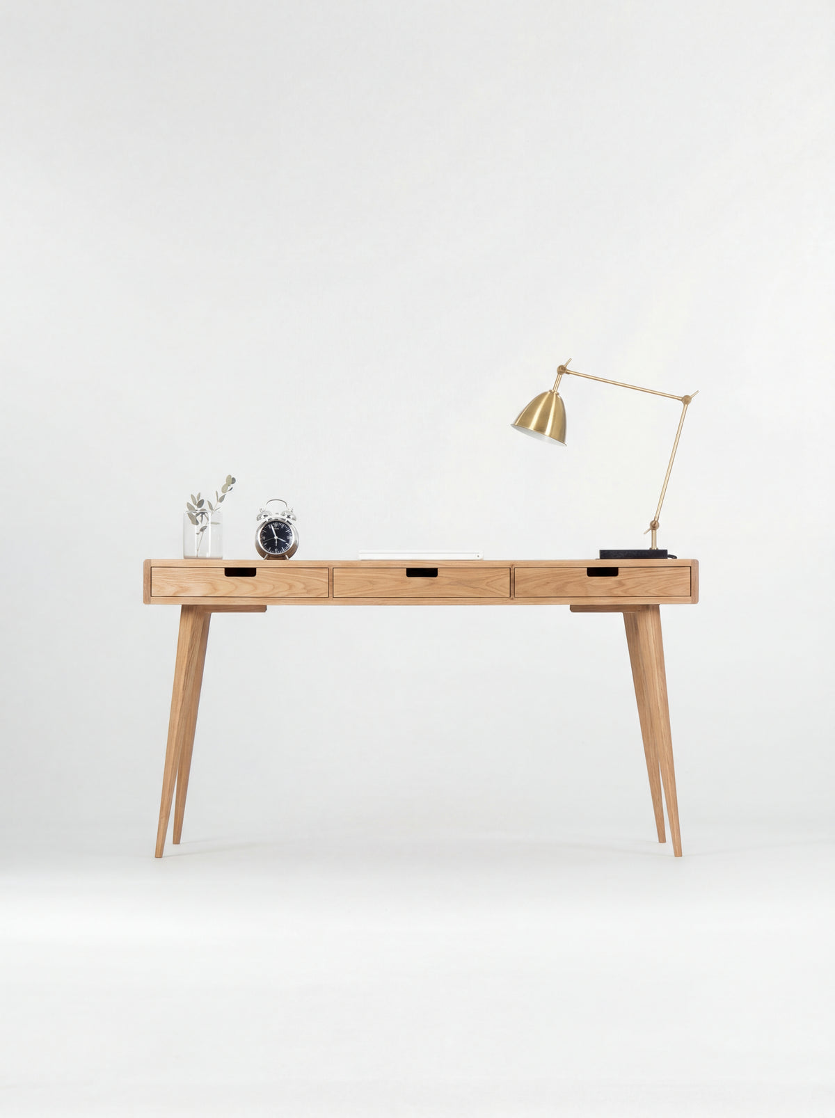 Front view of the HEIM 140 computer desk in oak wood, with three wooden drawer fronts, a brass lamp and alarm clock