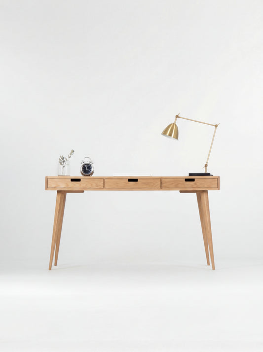 Front view of the HEIM 140 computer desk in oak wood, with three wooden drawer fronts, a brass lamp and alarm clock