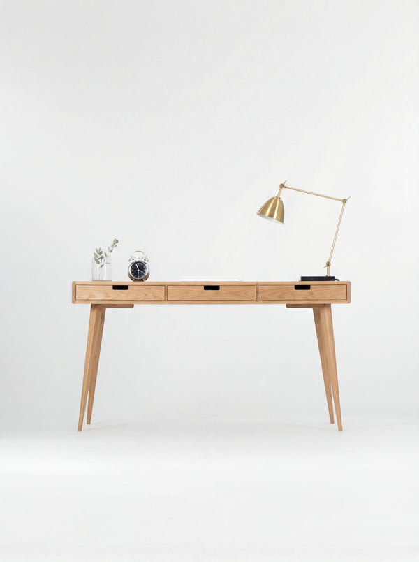 Front view of the HEIM 140 computer desk in oak wood, with three wooden drawer fronts, a brass lamp and alarm clock