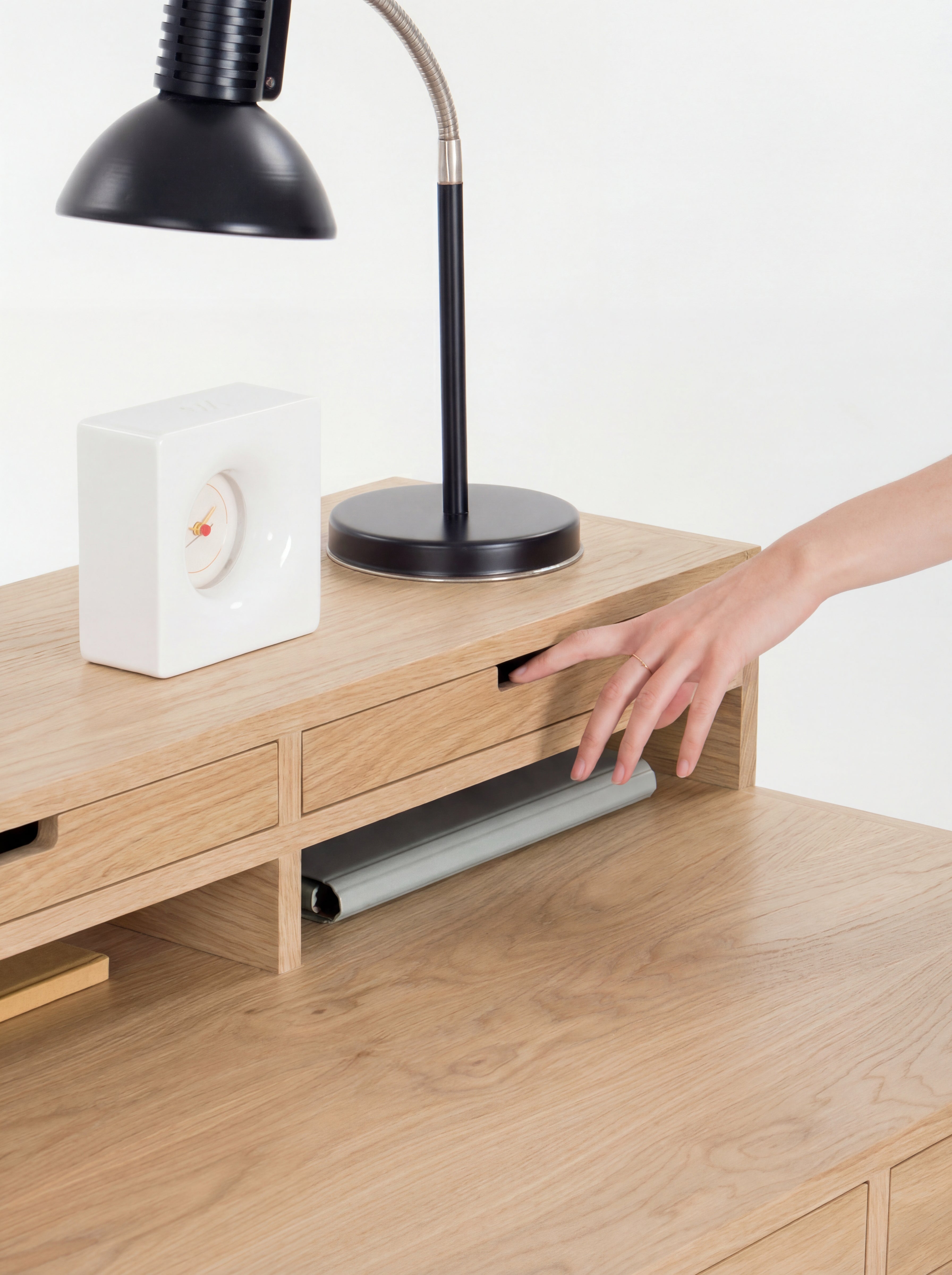 Hand opening a small drawer on a wooden desk with a lamp and clock on top.