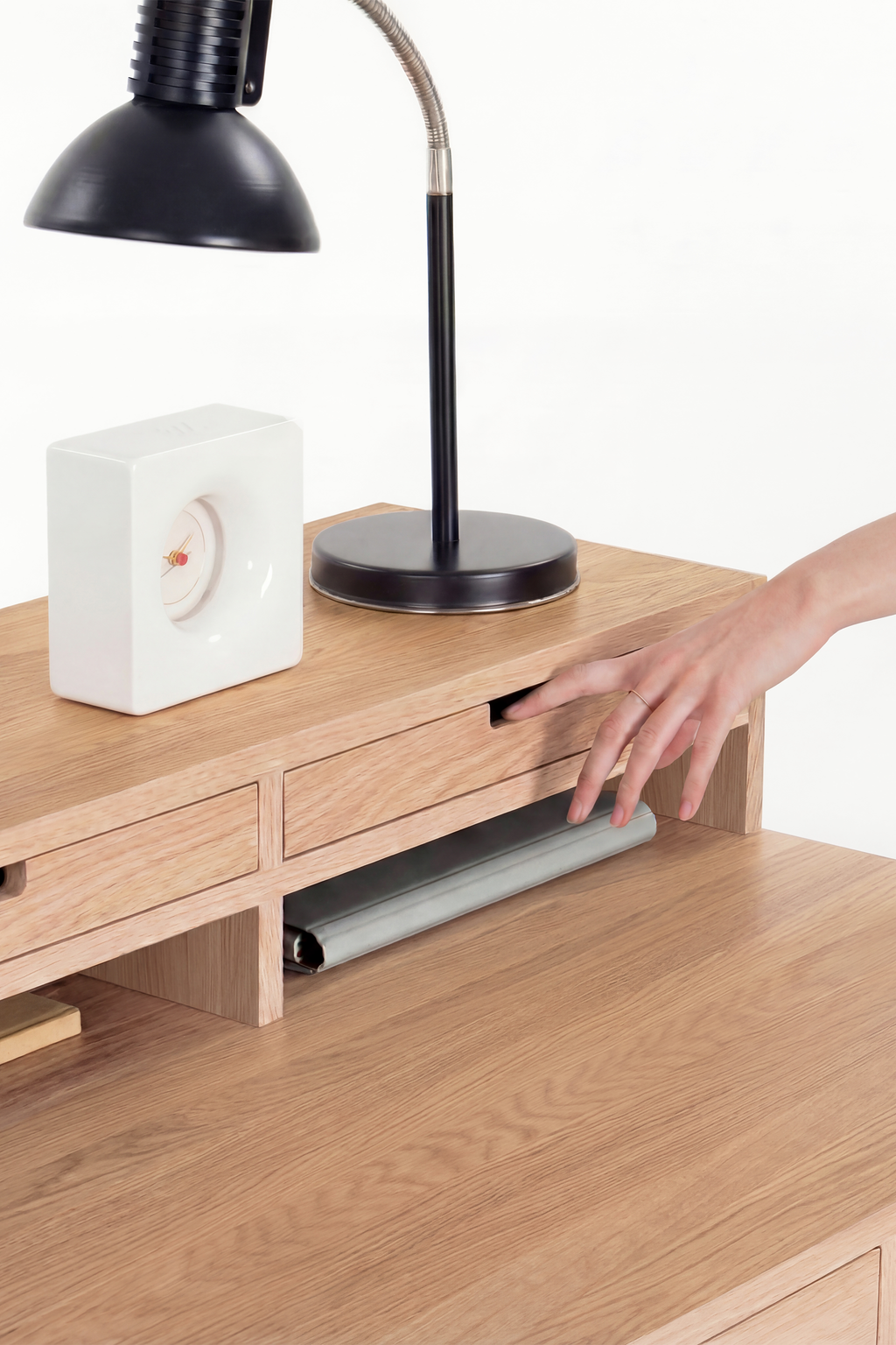 A hand pulls open a hutch drawer on the HEIM 140 in solid oak, with a white clock and black lamp above
