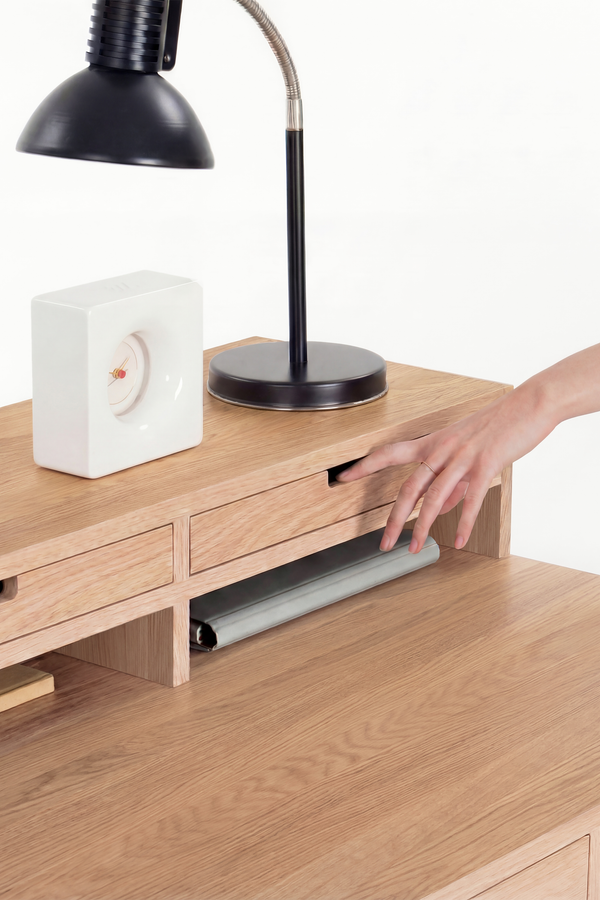 A hand pulls open a hutch drawer on the HEIM 140 in solid oak, with a white clock and black lamp above
