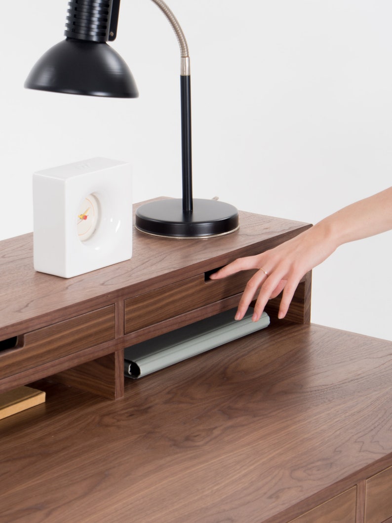 Hand opening a drawer on a wooden desk with a black lamp and white clock on top.