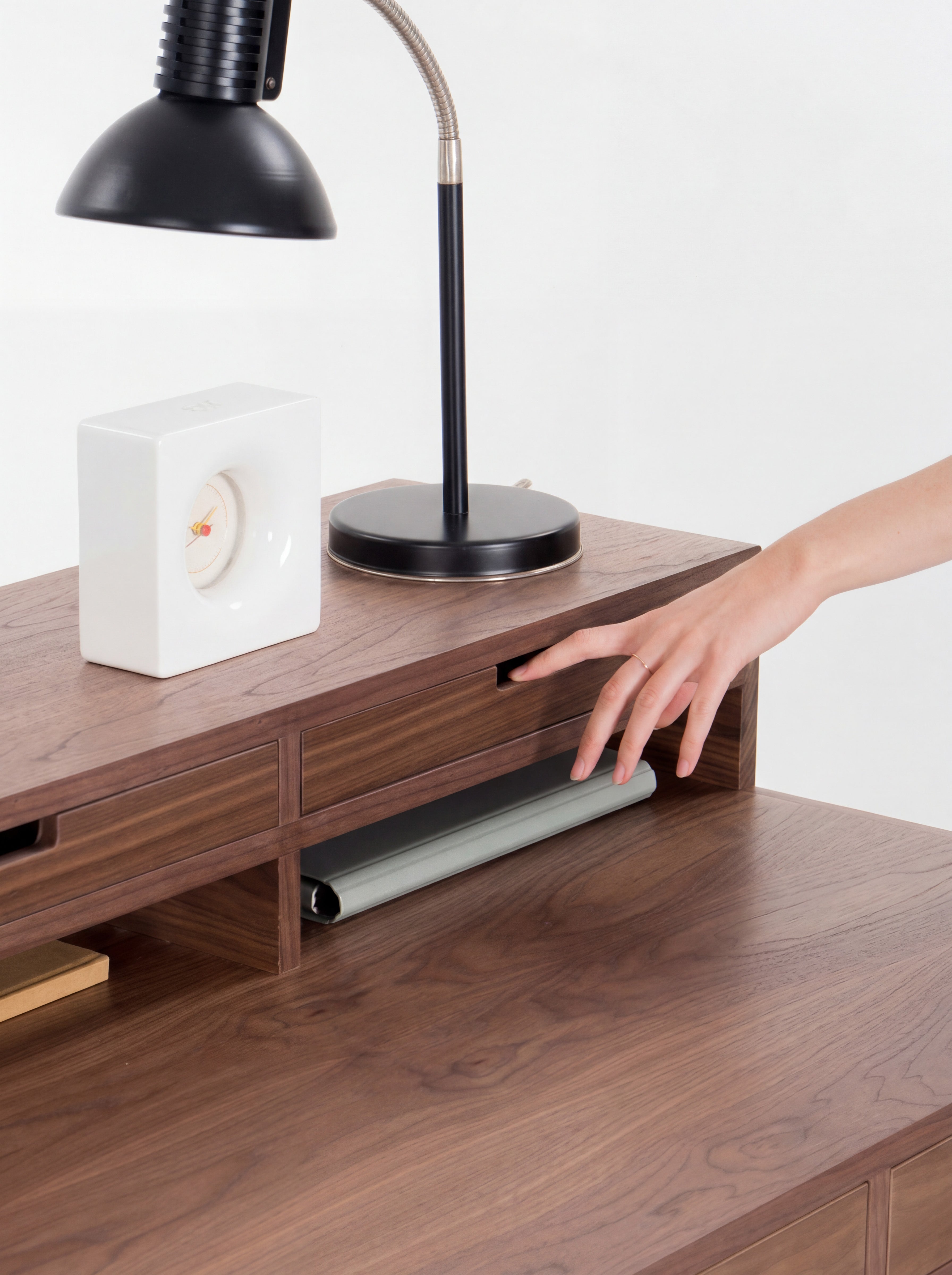 Hand opening a drawer on a wooden desk with a black lamp and white clock on top.