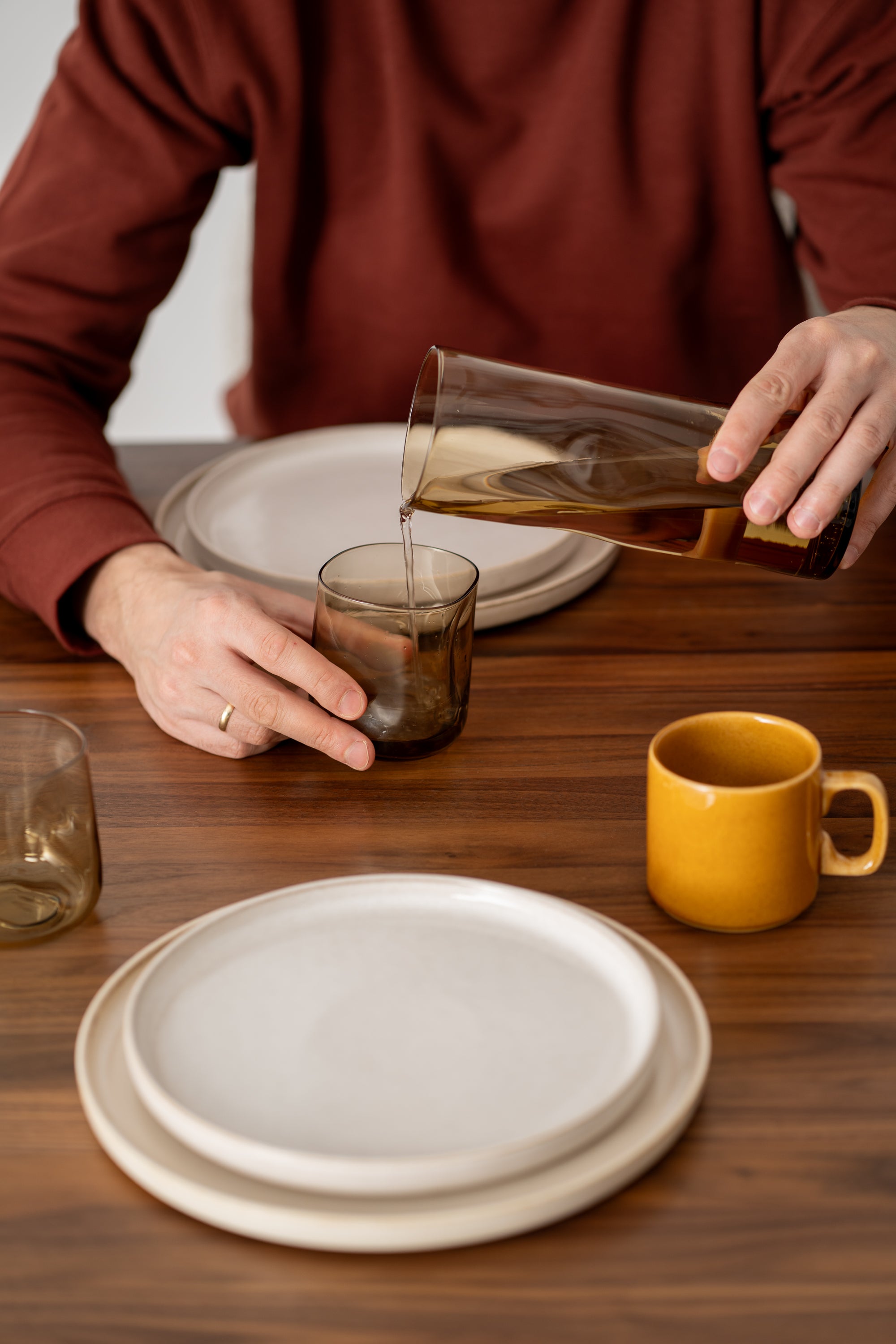 Person pouring liquid from a glass pitcher into a tumbler at a table set with plates and a yellow mug.