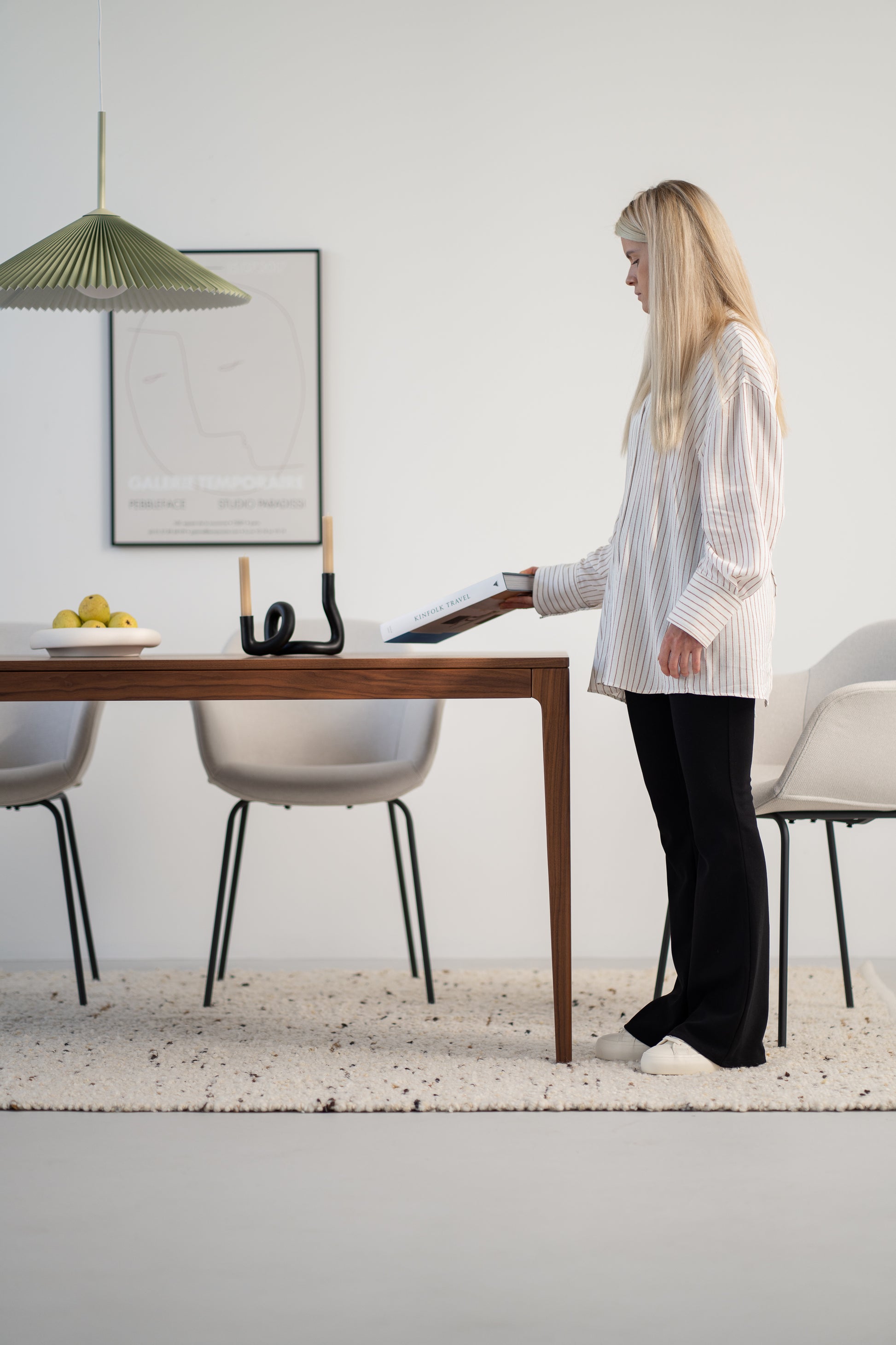 Woman in a striped blouse holding a book, standing by a dining table with chairs.