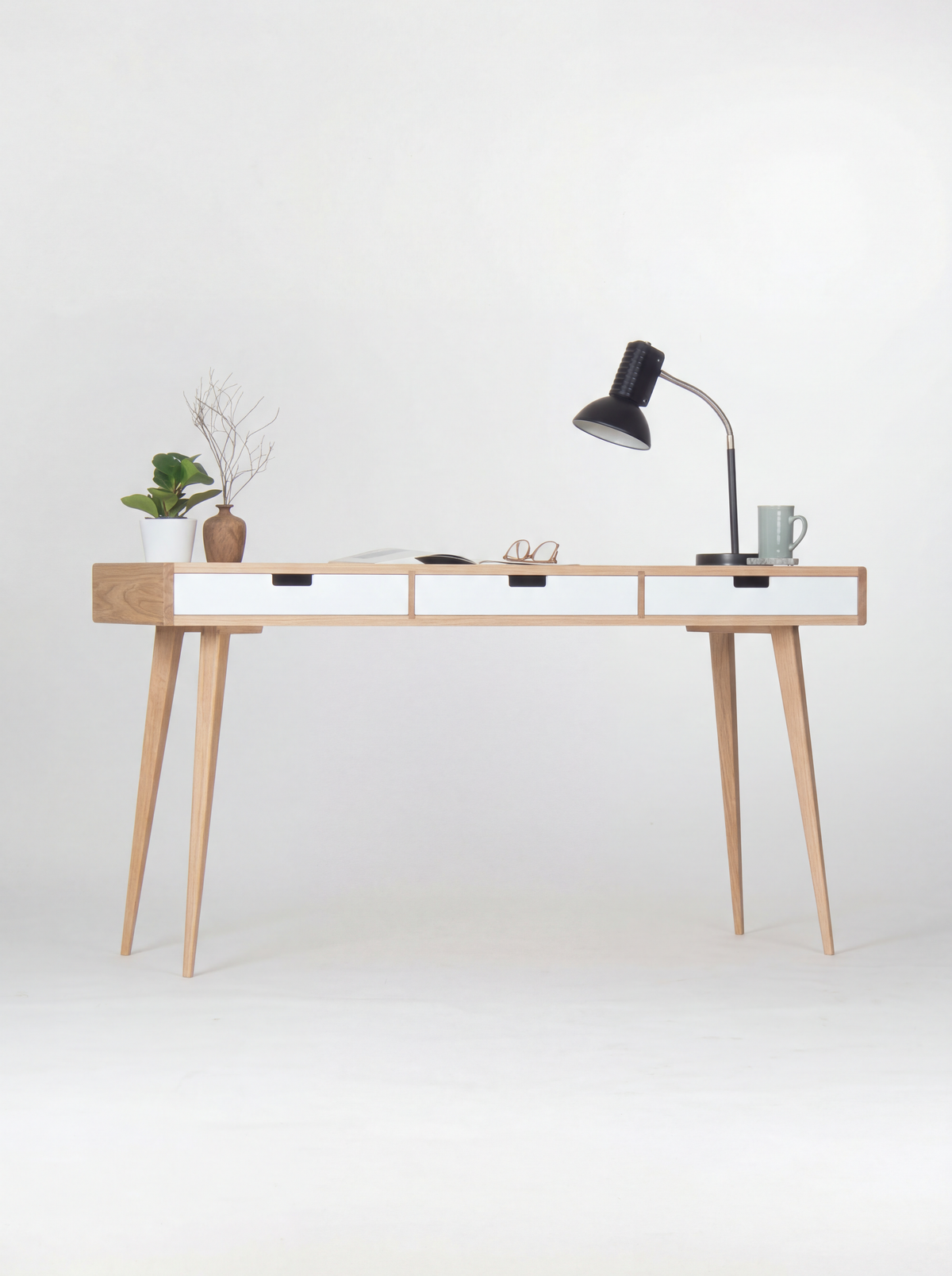 HEIM 140 desk in oak wood, full view with white drawer fronts, splayed legs, a black lamp and mug