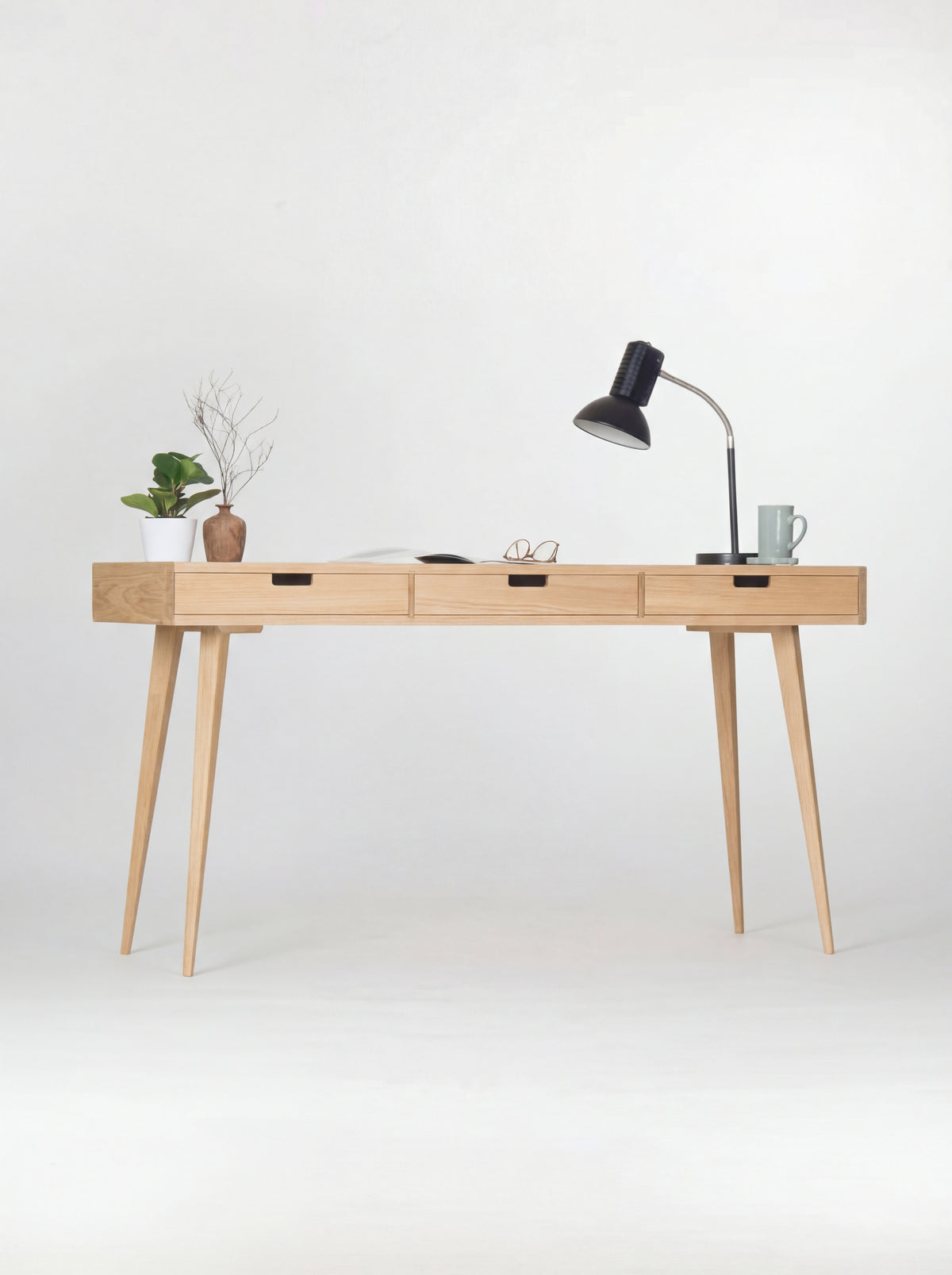 HEIM 140 desk in oak wood, straight-on view with three natural oak drawer fronts, a black lamp, plant and mug