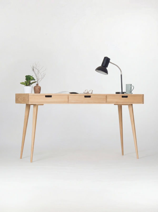 HEIM 140 desk in oak wood, straight-on view with three natural oak drawer fronts, a black lamp, plant and mug