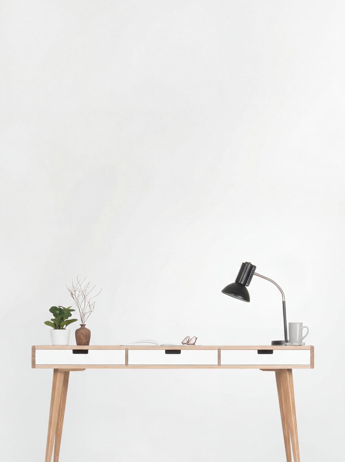 HEIM 140 desk in oak wood against a white wall, with three white drawer fronts, a black lamp, plant and mug