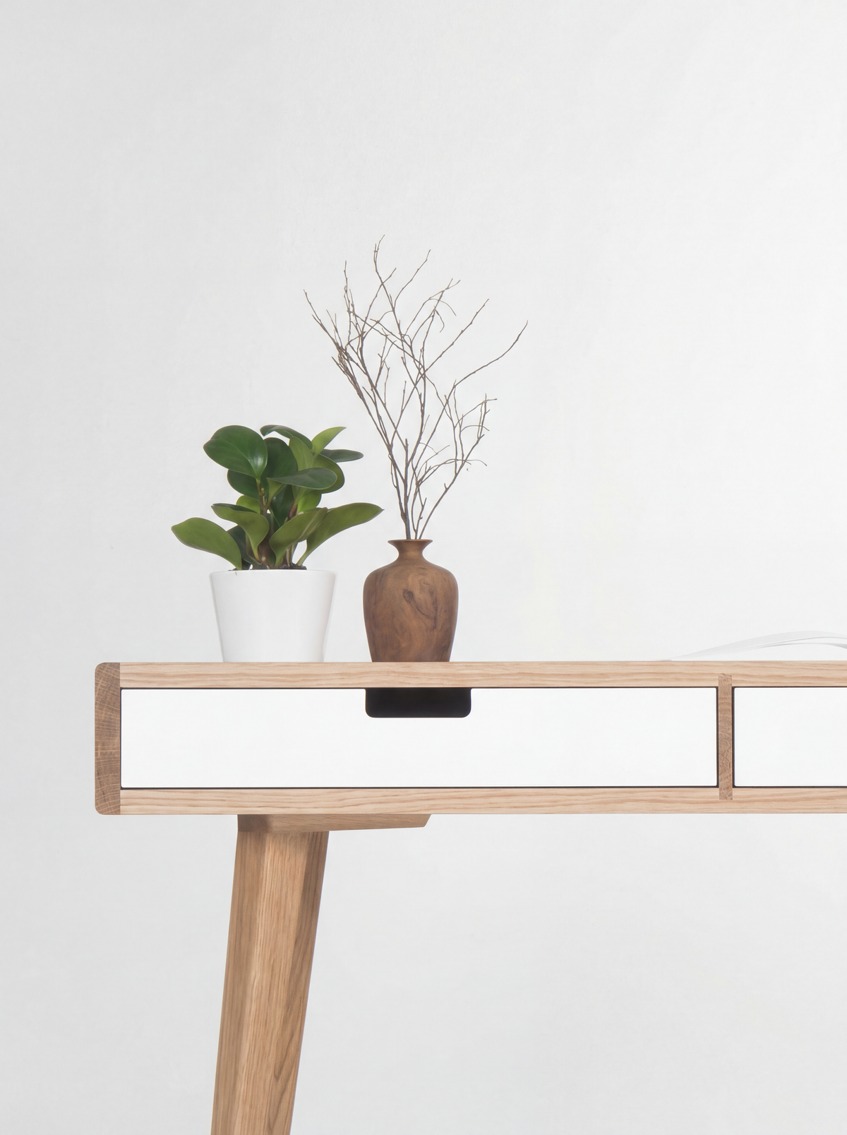 Close-up of the HEIM 140 desk in oak wood, with a white drawer front, cut-out grip, oak leg and a vase with twigs