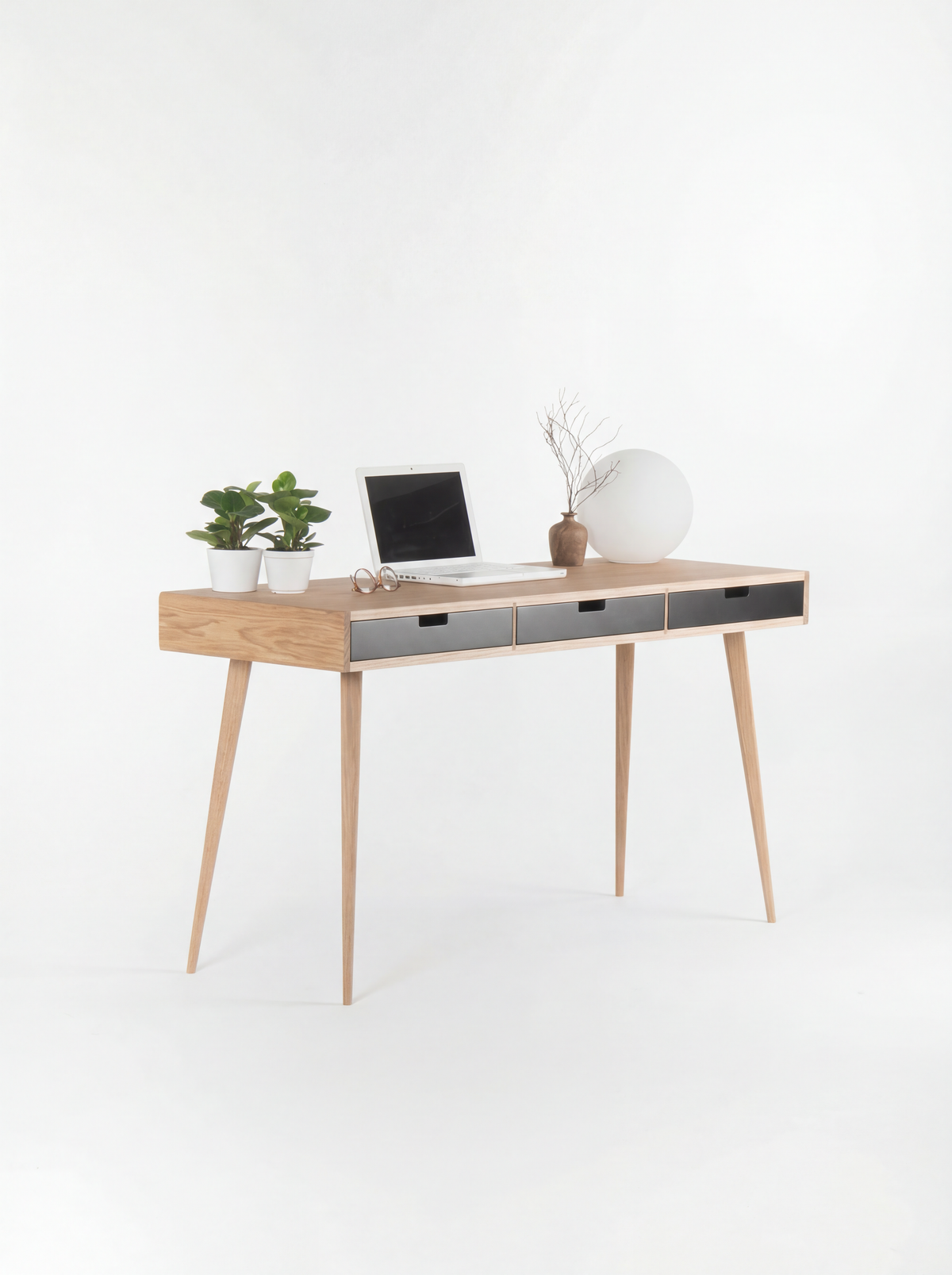 Three-quarter view of the HEIM 140 desk in oak wood, with its oak side, black drawer fronts, laptop and globe lamp